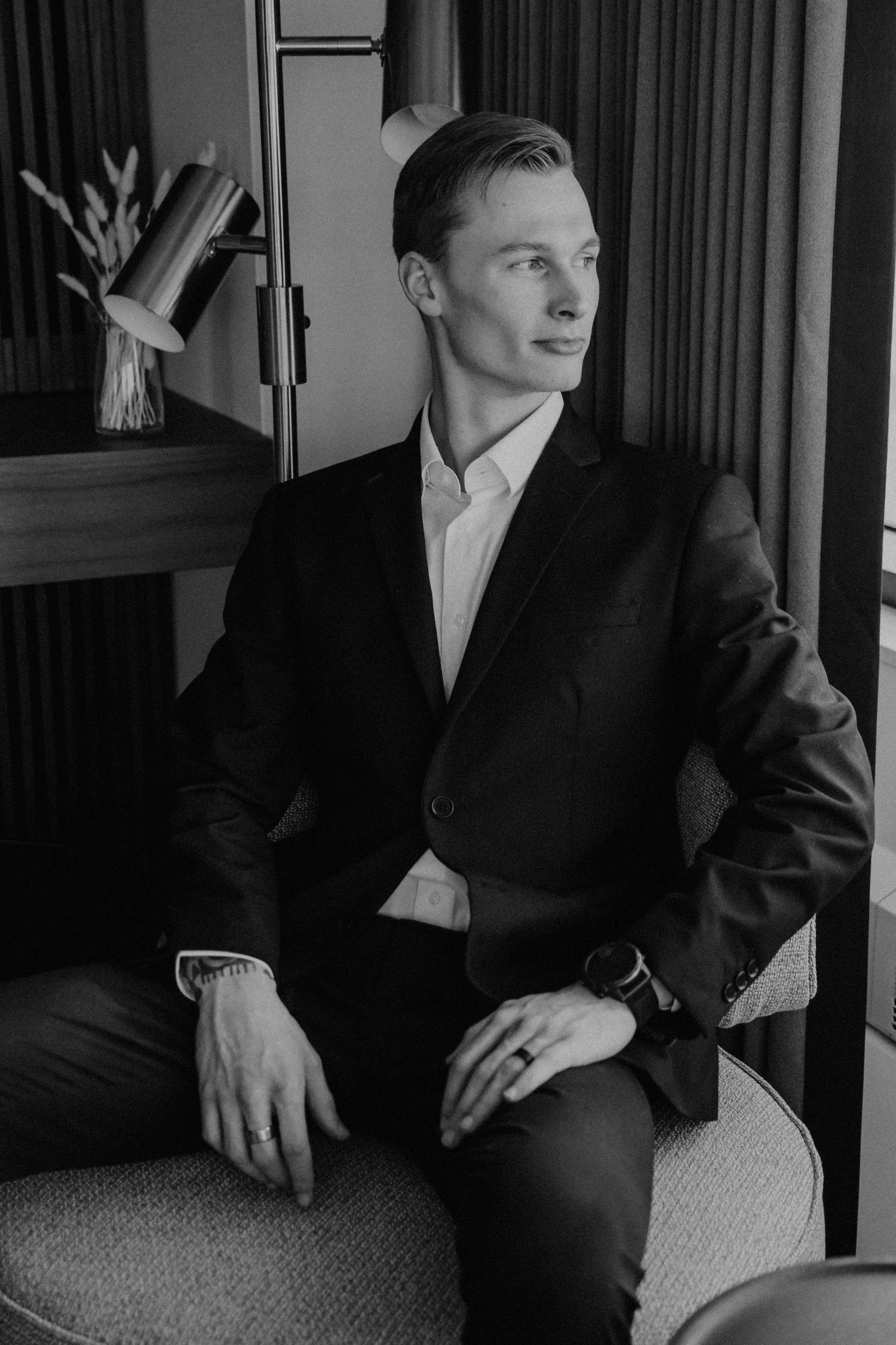 Black and white portrait of elegant groom in black suit seated in stylish hotel room.