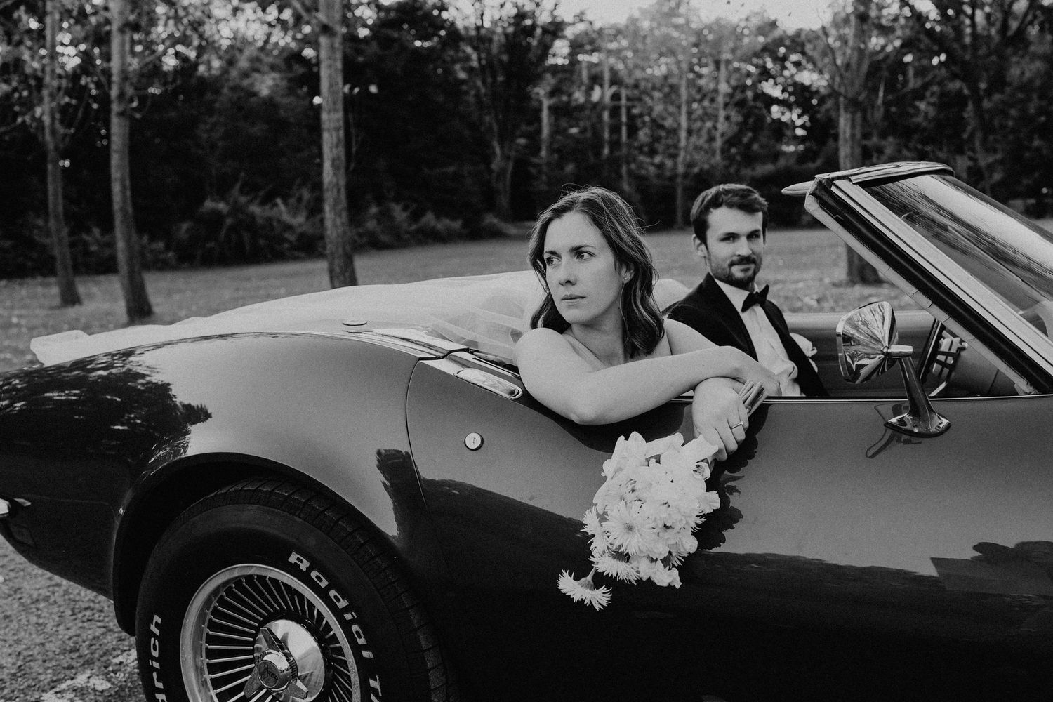 Bride leaning over door of classic black Corvette convertible with groom, dramatic black and white wedding photo.