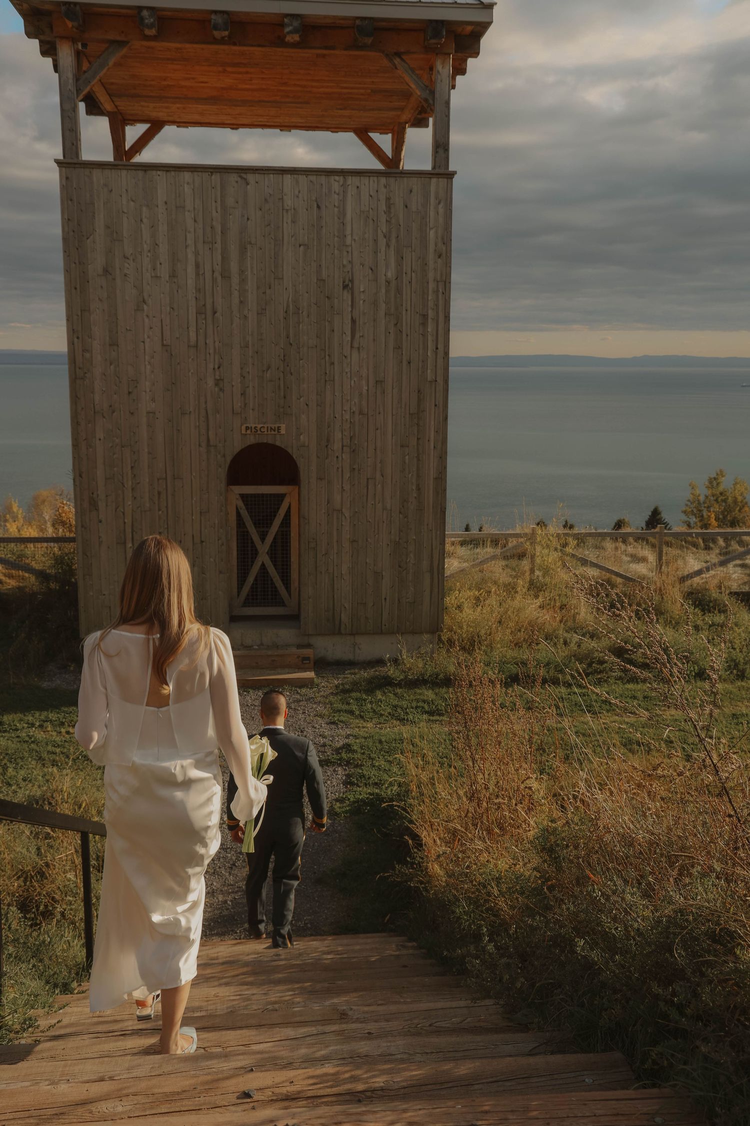 Bride in white dress and groom walking hand in hand toward wooden tower overlooking the sea.