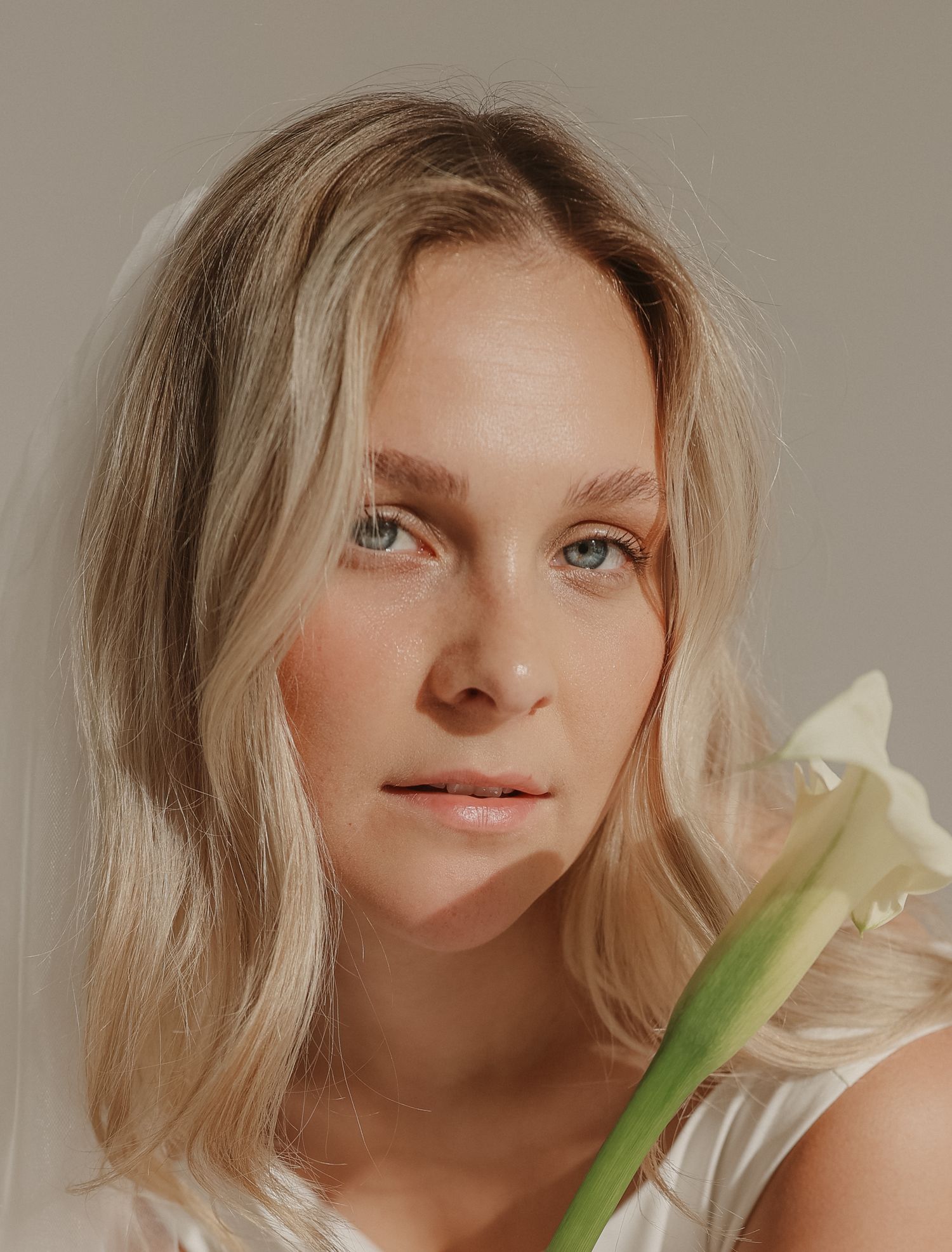 Blonde woman with blue eyes holding a white calla lily flower in a minimalist portrait.