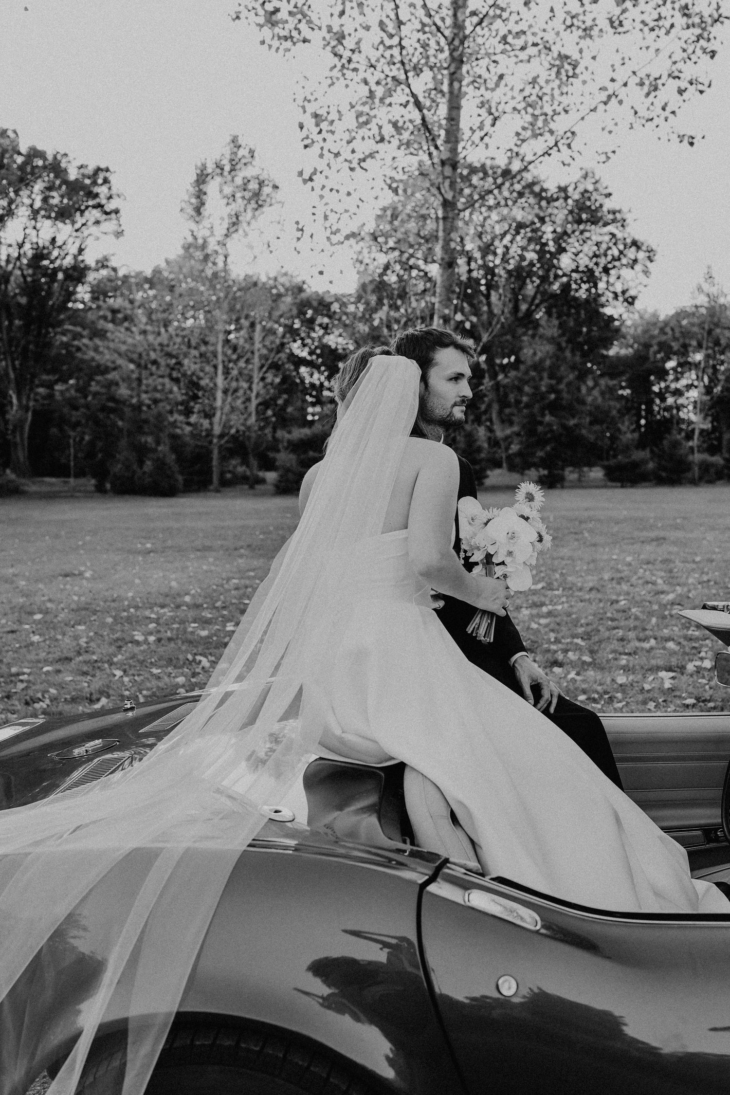 Bride in flowing veil sits on vintage convertible with groom in autumn park, black and white wedding photo.