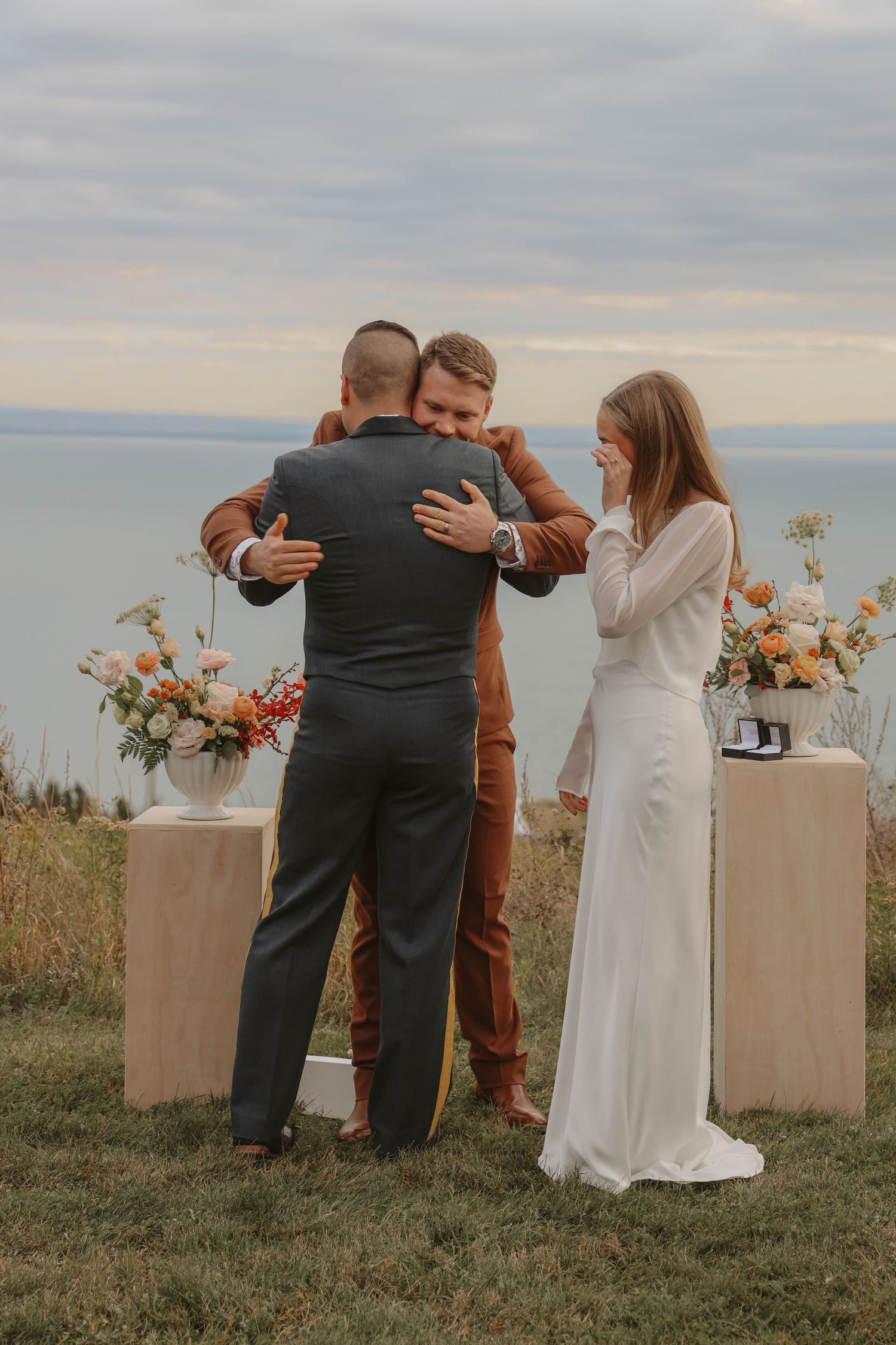Groom embraces guest at outdoor cliffside wedding ceremony while emotional bride looks on.