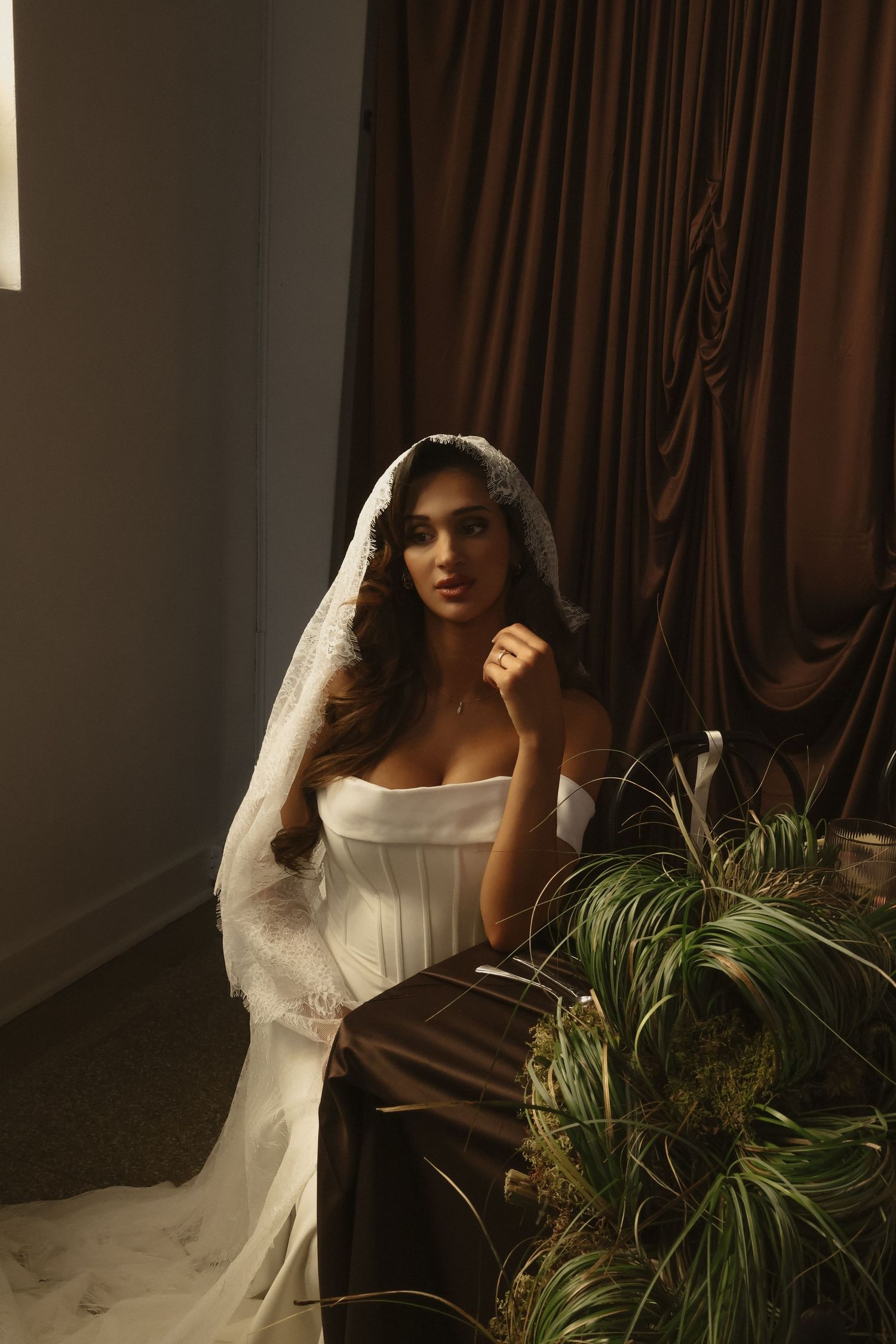 A bride in a white off-shoulder gown and lace veil sits elegantly beside green plants against rich brown drapes.