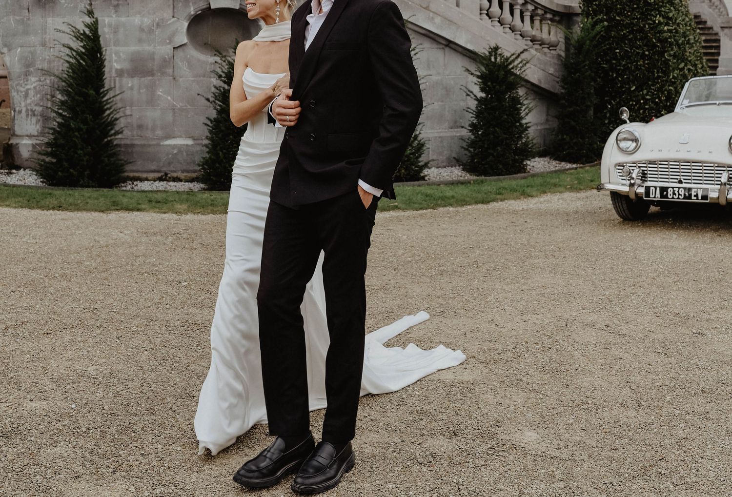 Bride and groom embrace in front of a vintage white car at an elegant chateau wedding venue.