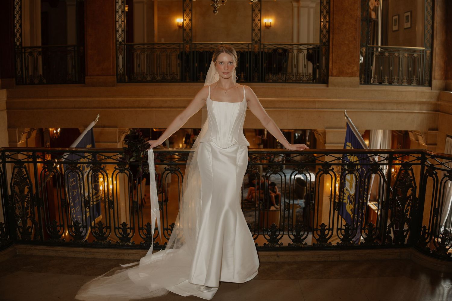 Elegant bride in a white satin mermaid wedding gown with train, posing on ornate balcony stairs.
