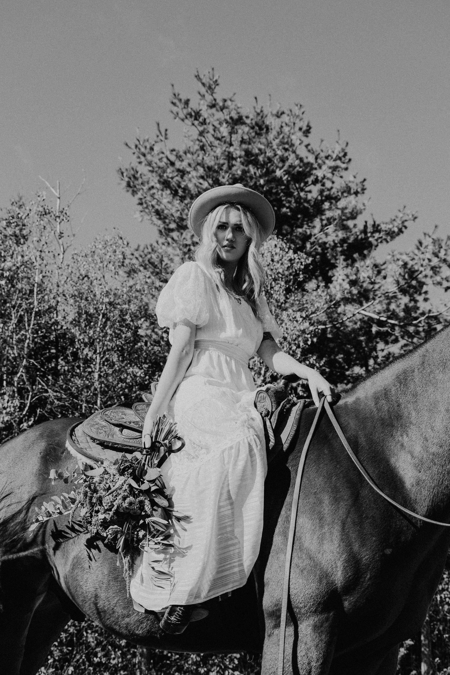 Black and white photo of a boho bride in a white dress and hat posed on horseback outdoors.