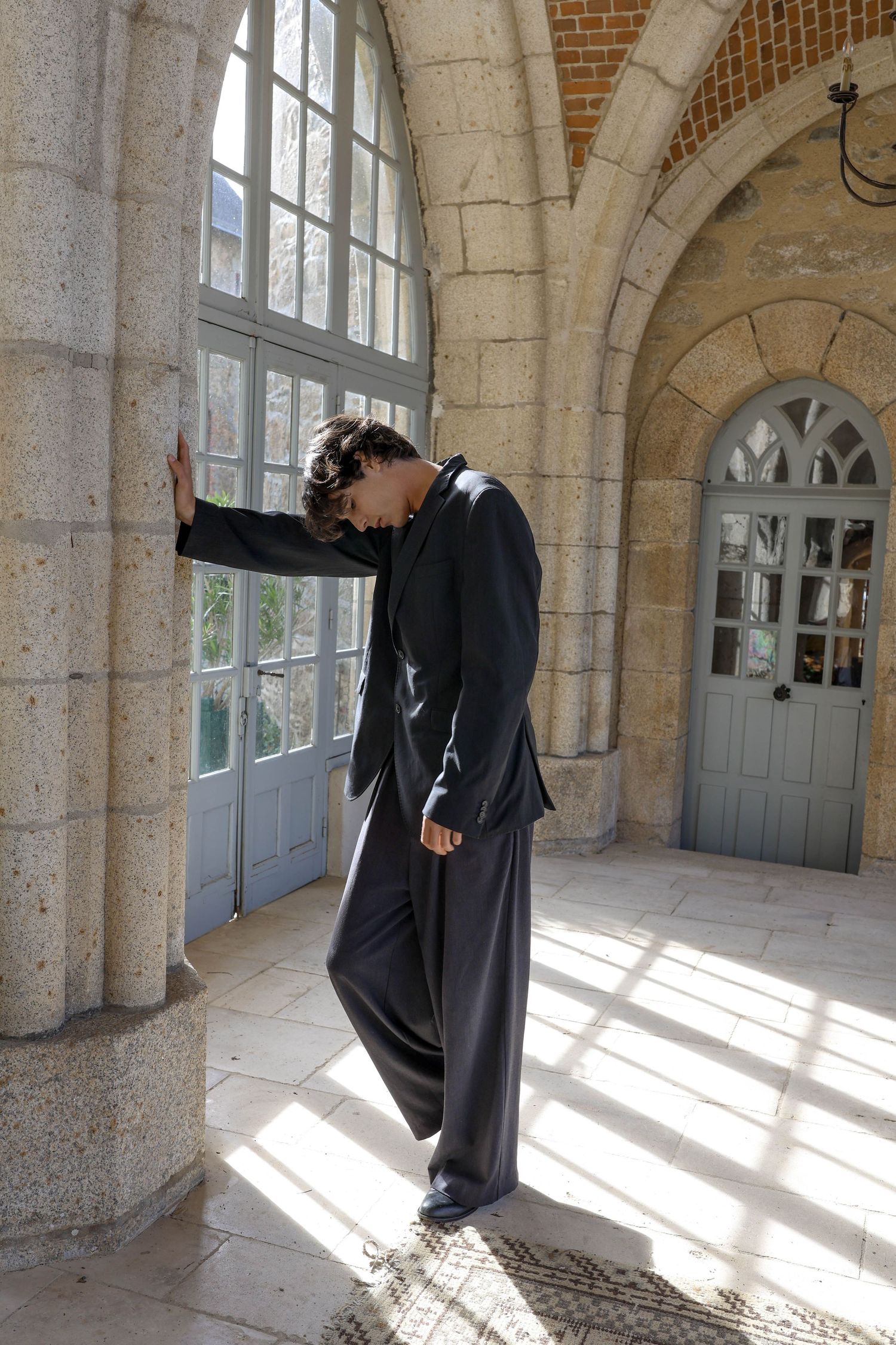 Person in dark oversized suit leaning against arched stone window in historic building.