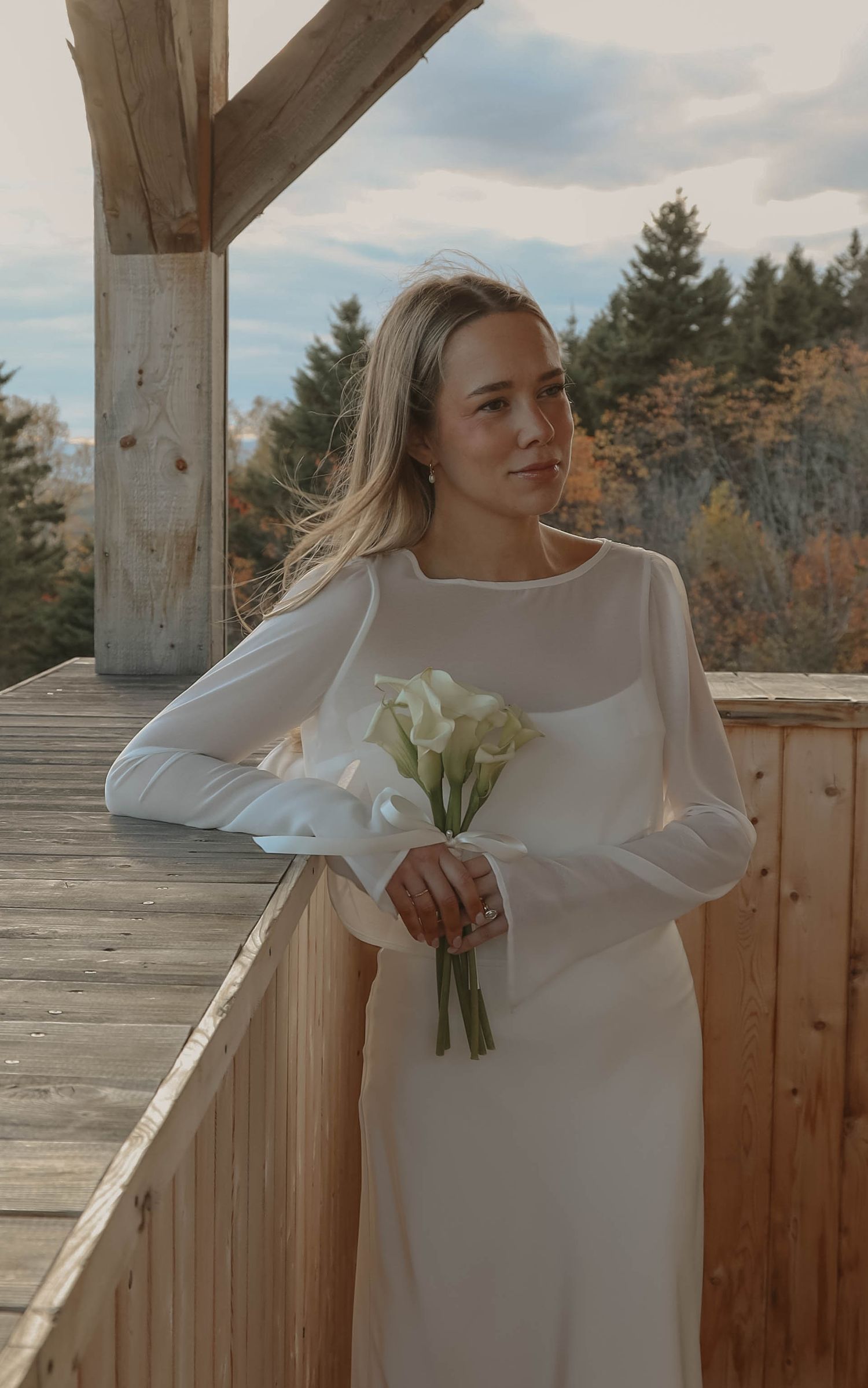 Bride in minimalist long-sleeve white gown holding calla lily bouquet on wooden deck outdoors.
