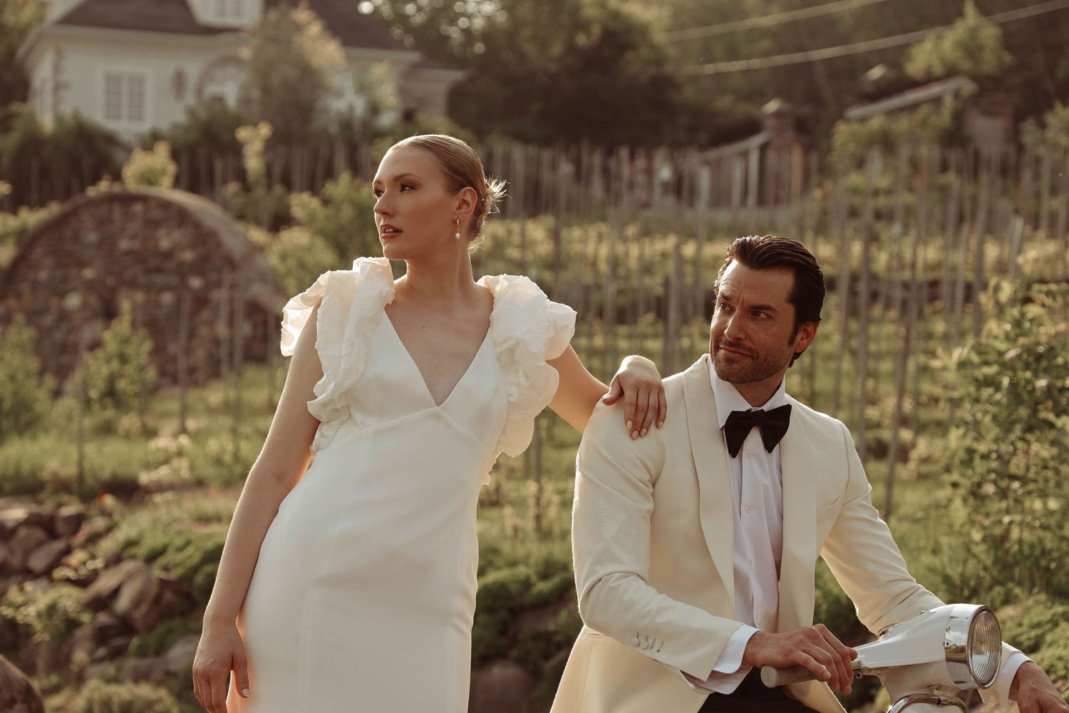 Bride in white V-neck gown and groom in white tuxedo posing together at vineyard wedding.