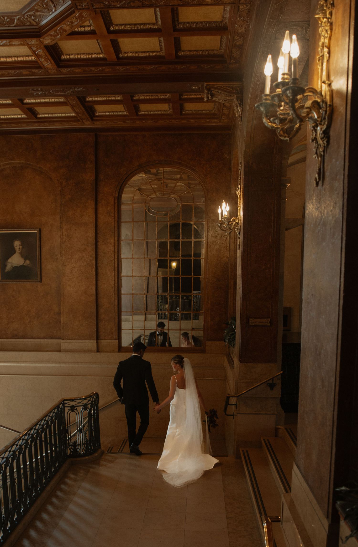 Bride and groom holding hands walking down grand staircase in ornate golden ballroom with elegant chandeliers.