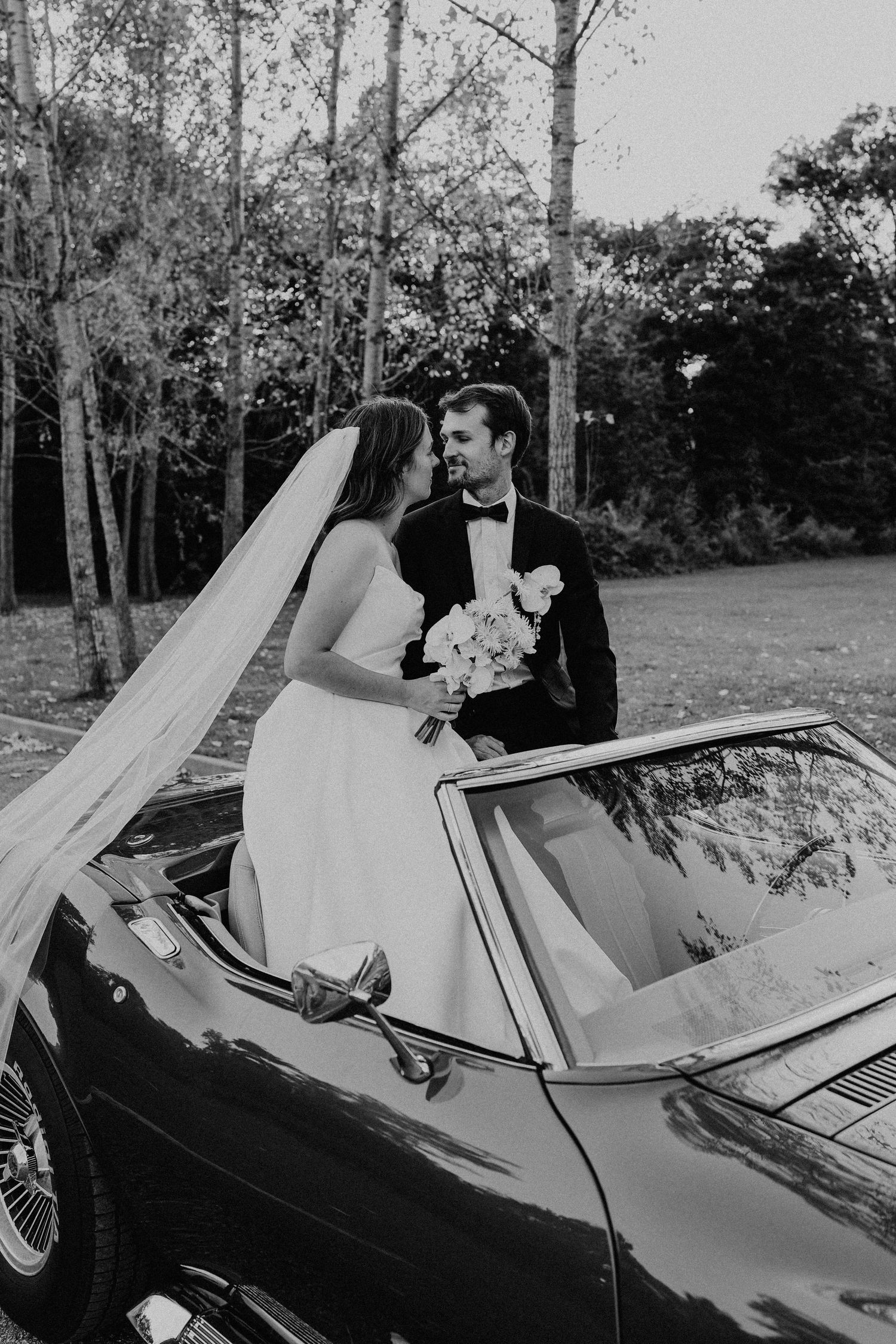 Bride and groom posing beside vintage convertible car in park setting, black and white wedding photography.