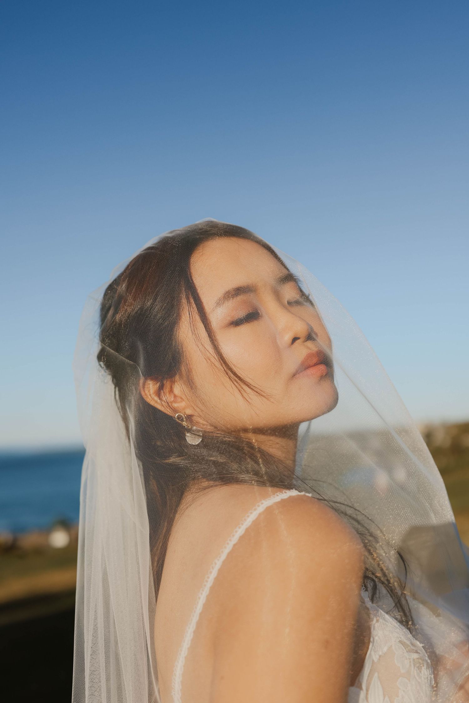 Beautiful Asian bride wearing a delicate veil poses against a clear blue sky near the ocean.