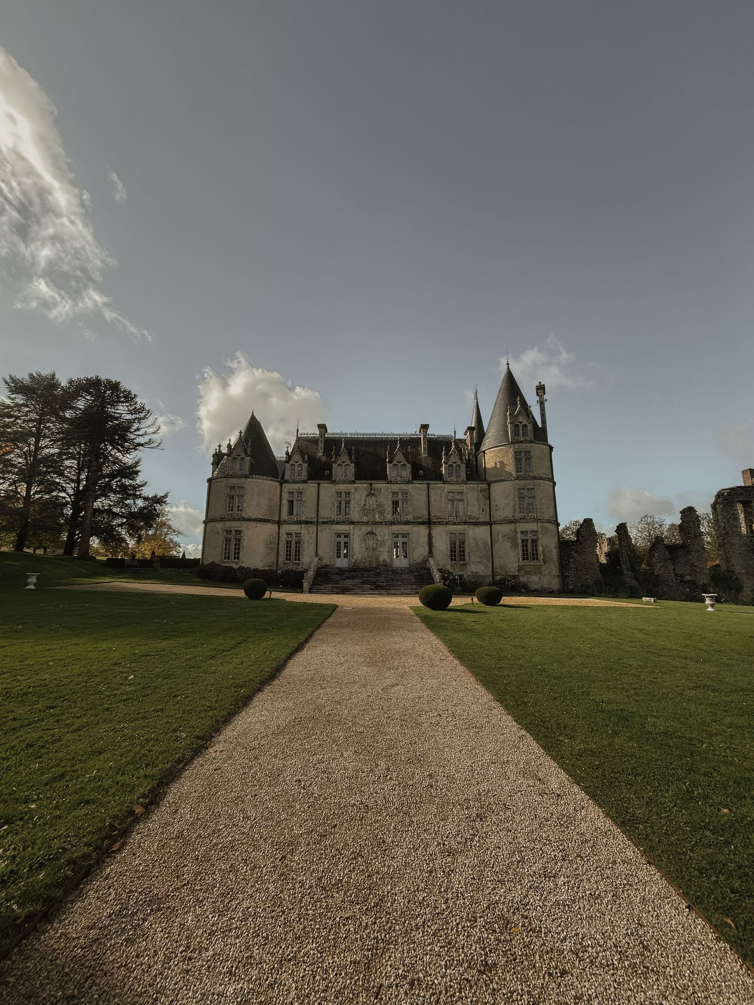 Grand French chateau with manicured grounds and gravel pathway under dramatic cloudy sky.