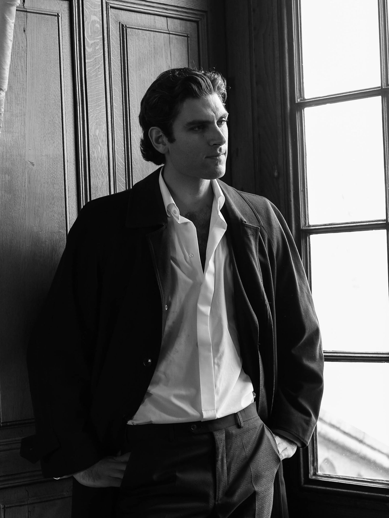 Stylish man in dark blazer and white shirt leaning against wooden wall by window in black and white portrait.
