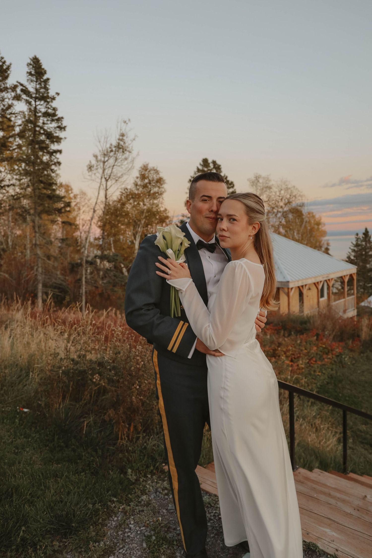 Bride and groom embracing at sunset, groom in military dress uniform holding white calla lilies.