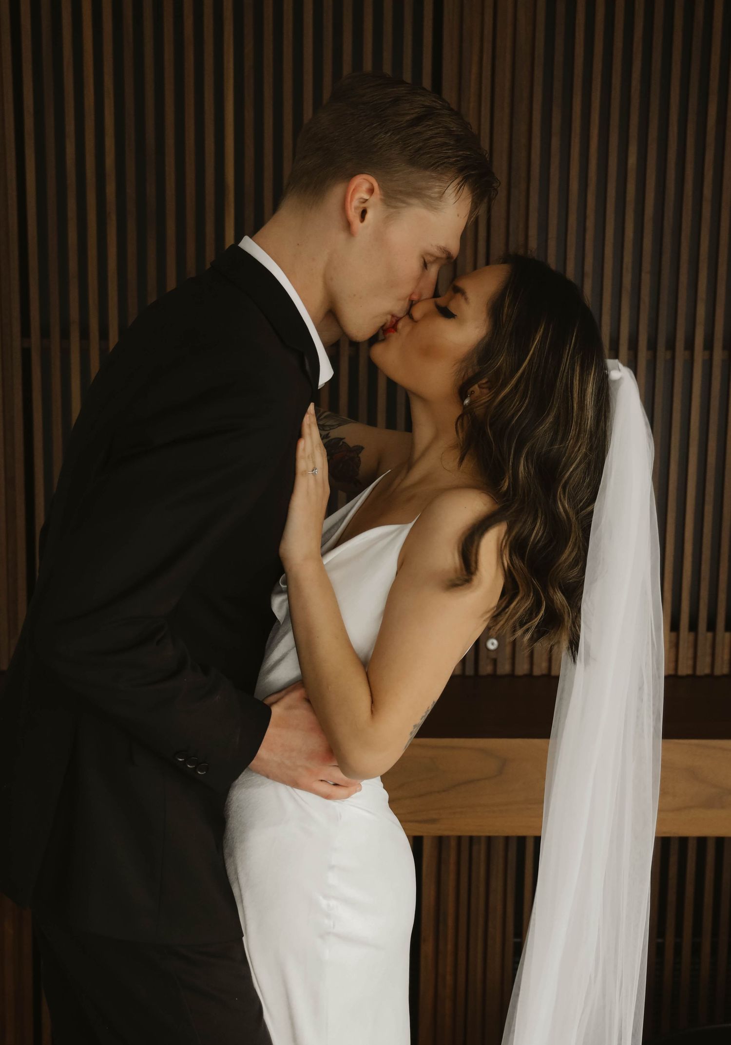 Bride in white gown with veil kissing groom in black suit against wooden backdrop.