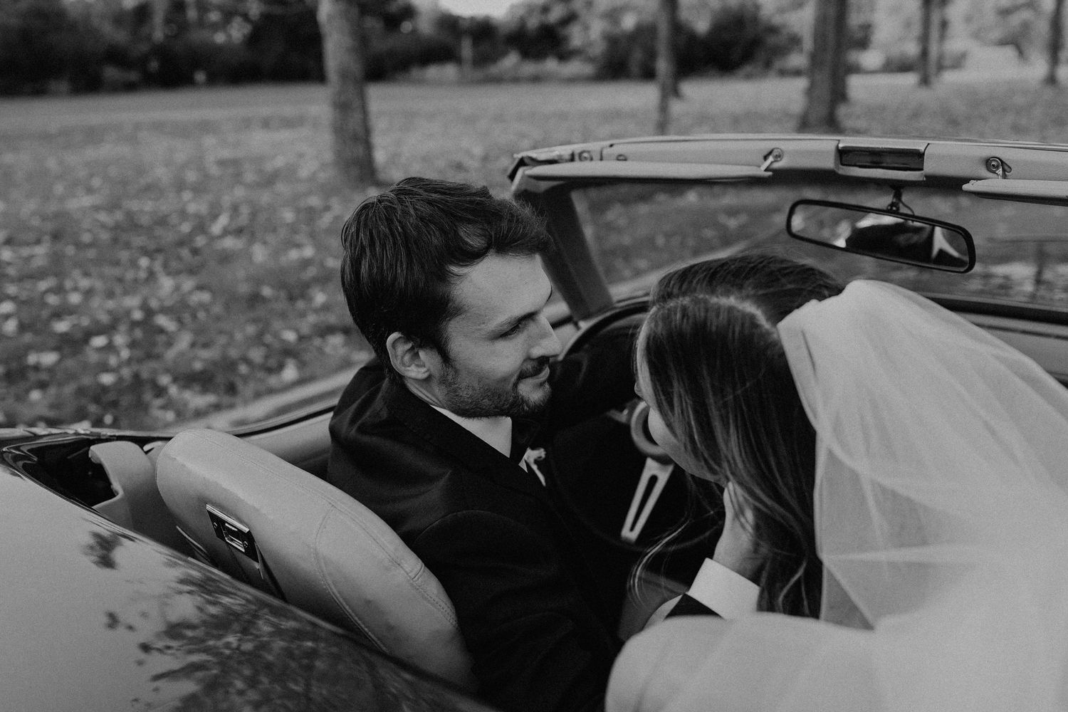 Black and white close-up of smiling bride and groom sharing intimate moment inside vintage convertible car.