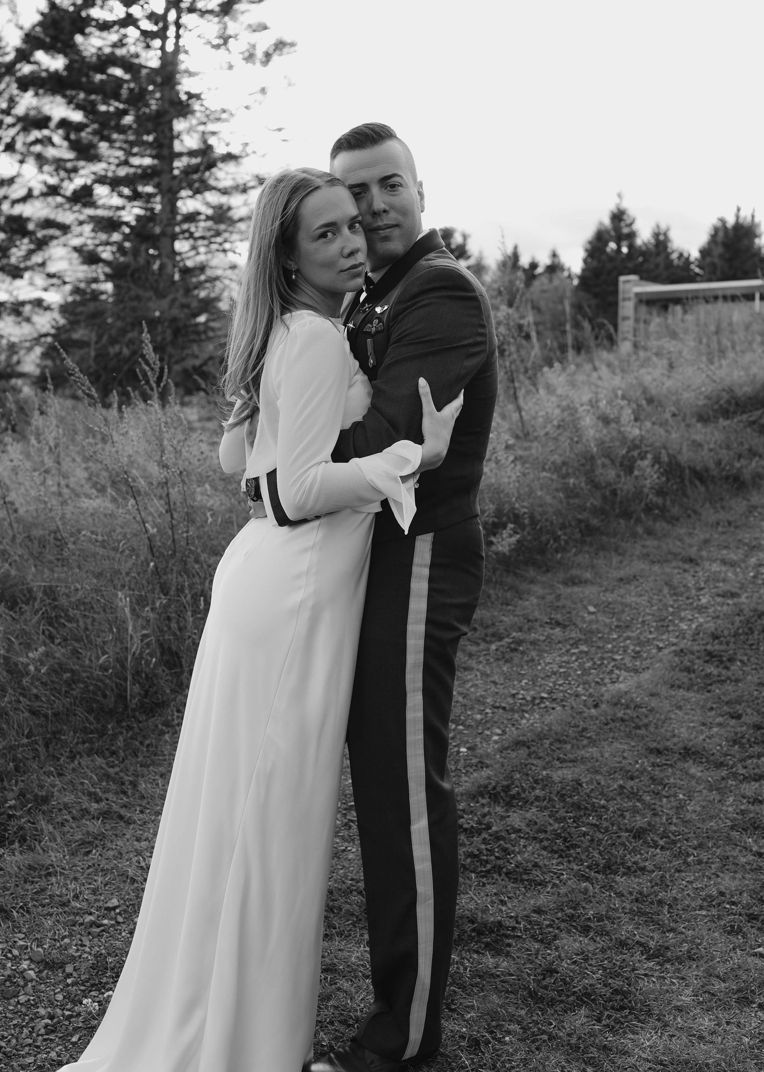 Black and white portrait of military groom and bride embracing outdoors surrounded by trees at dusk.