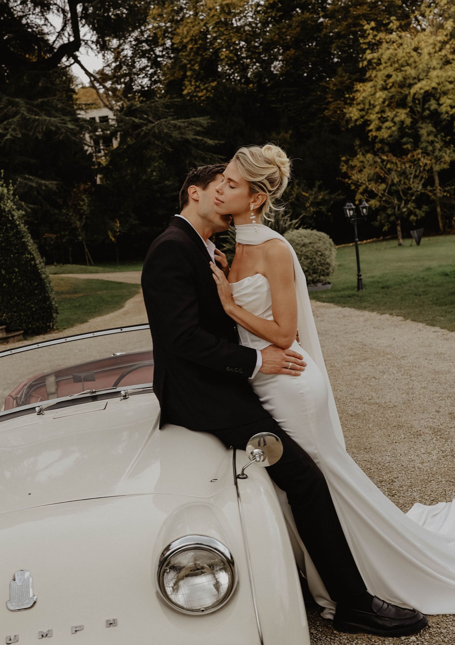Bride and groom share a romantic kiss while sitting on a classic white vintage car at chateau wedding.