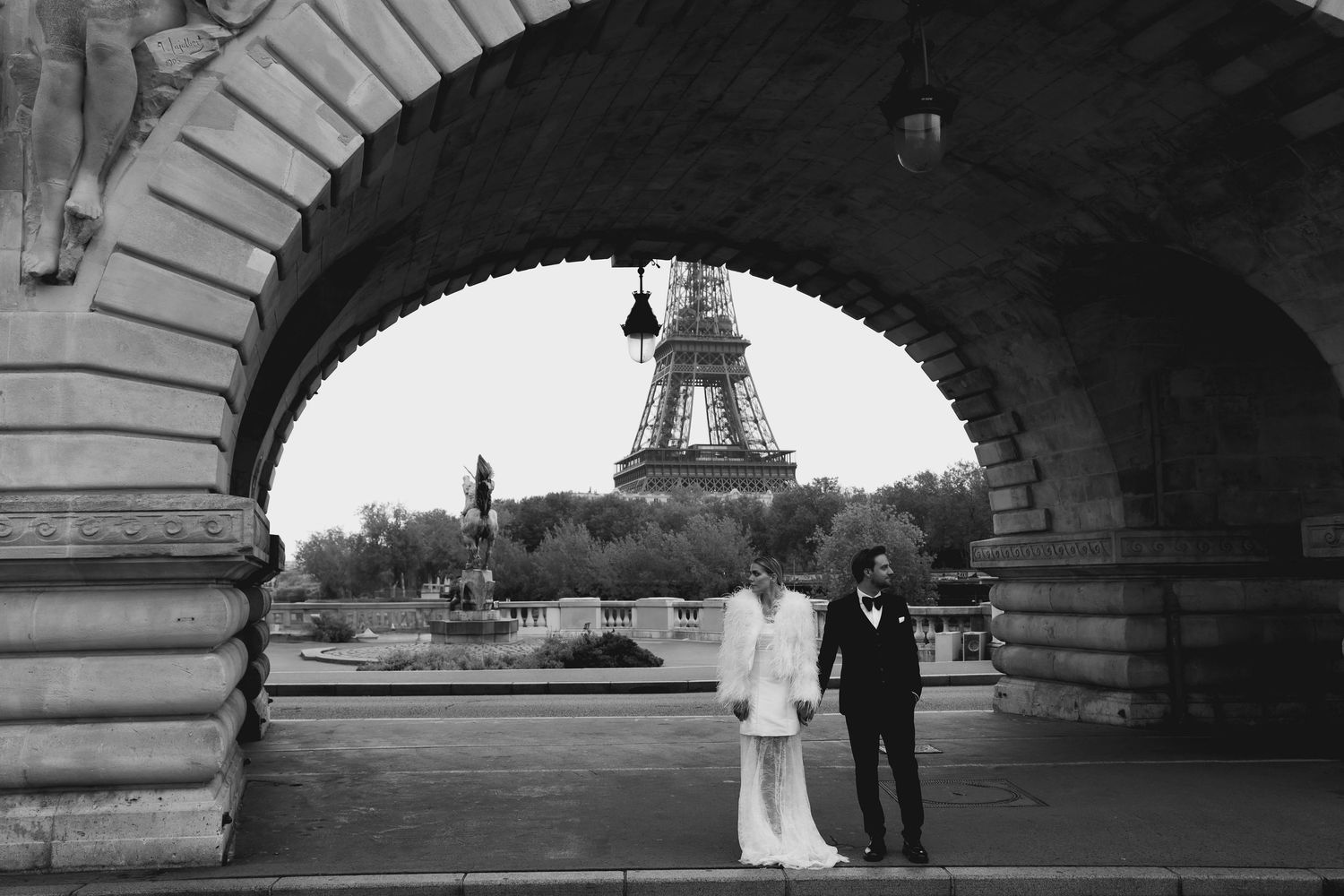 Black and white photo of bride and groom framed under a bridge arch with Eiffel Tower.