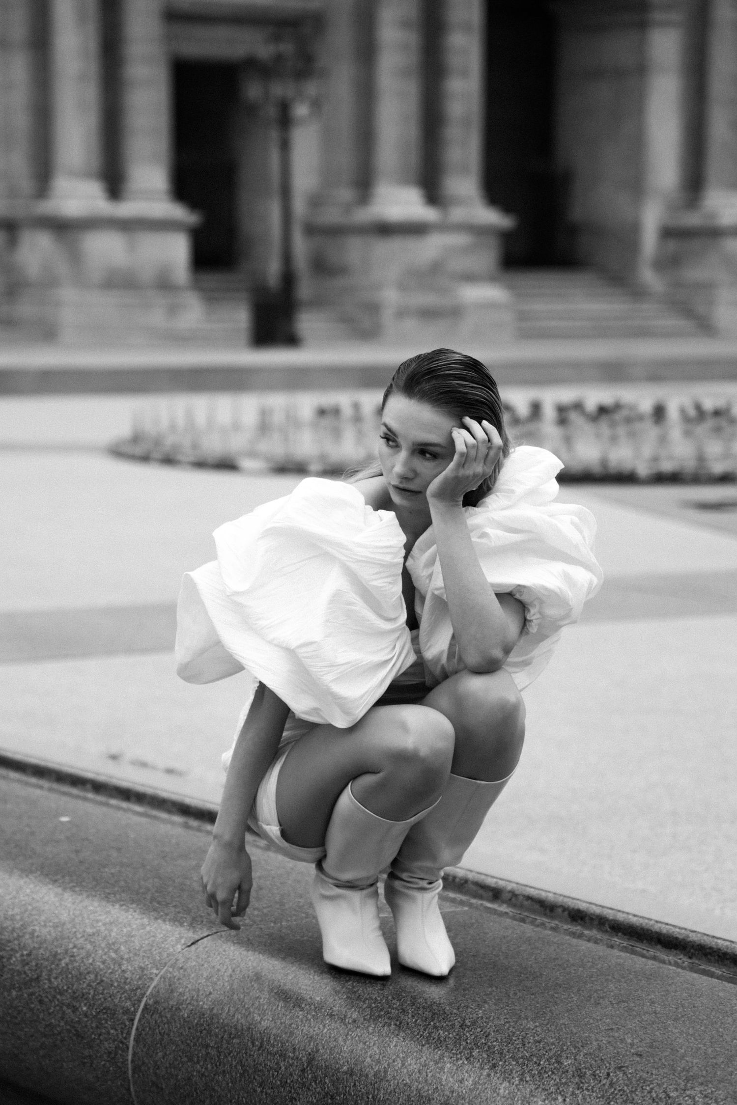 Black and white photo of woman in white ruffled dress crouching on Parisian street in white boots.