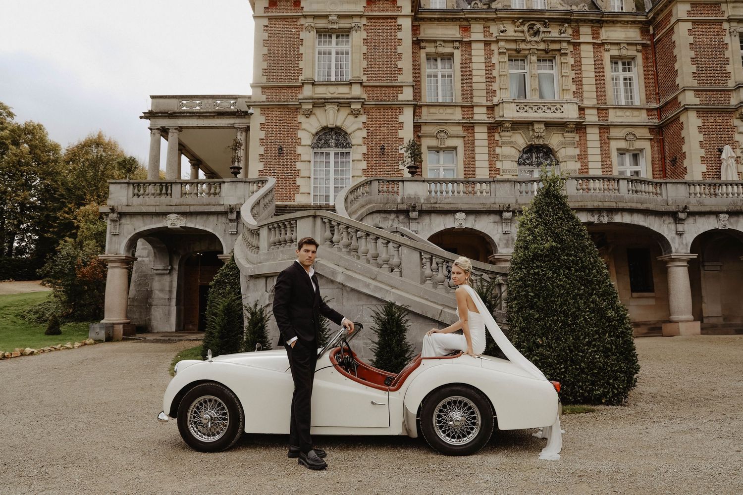 Bride and groom posing with vintage white car in front of elegant chateau wedding venue.