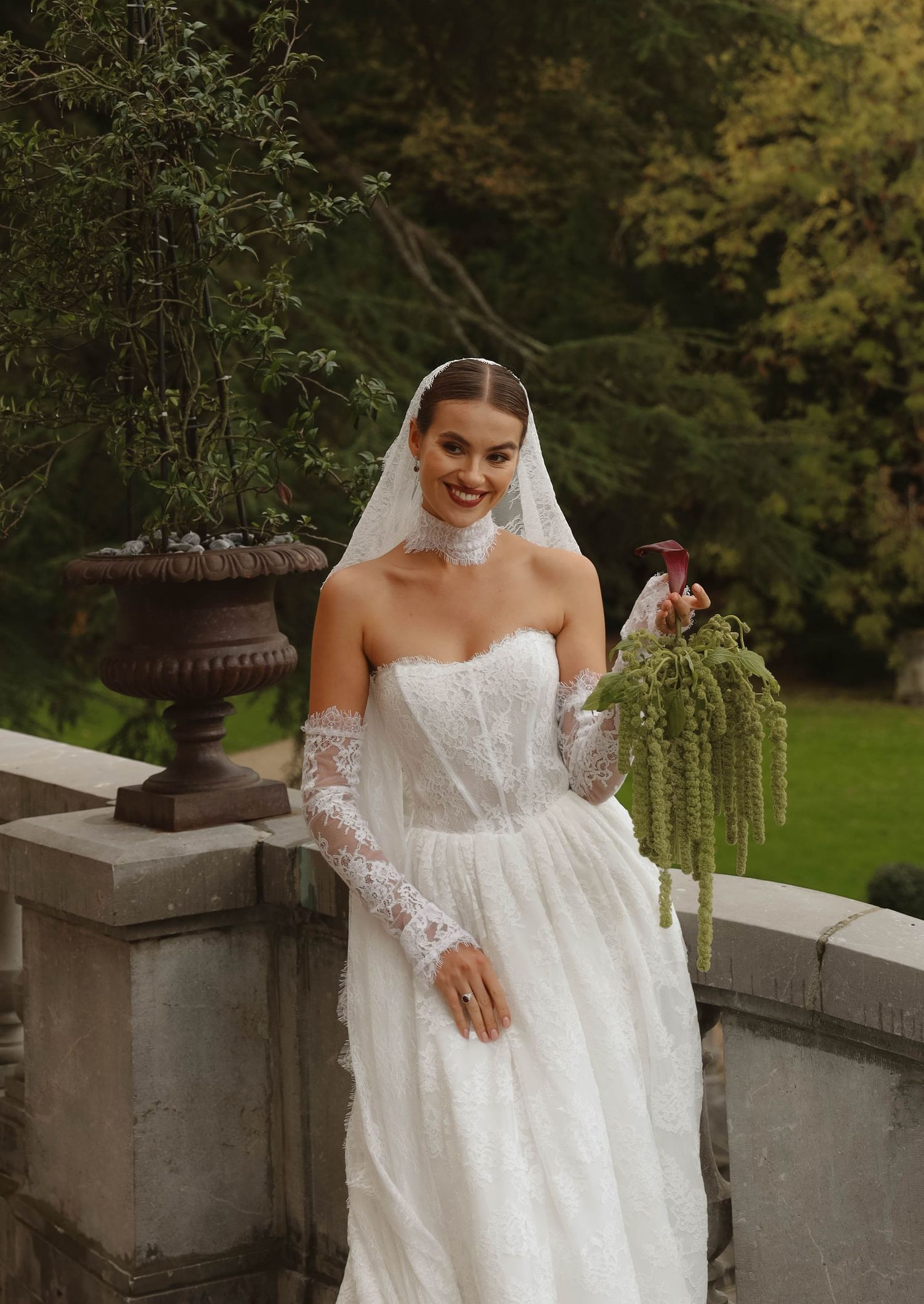 Smiling bride in strapless lace wedding dress with veil holding green floral bouquet in garden.