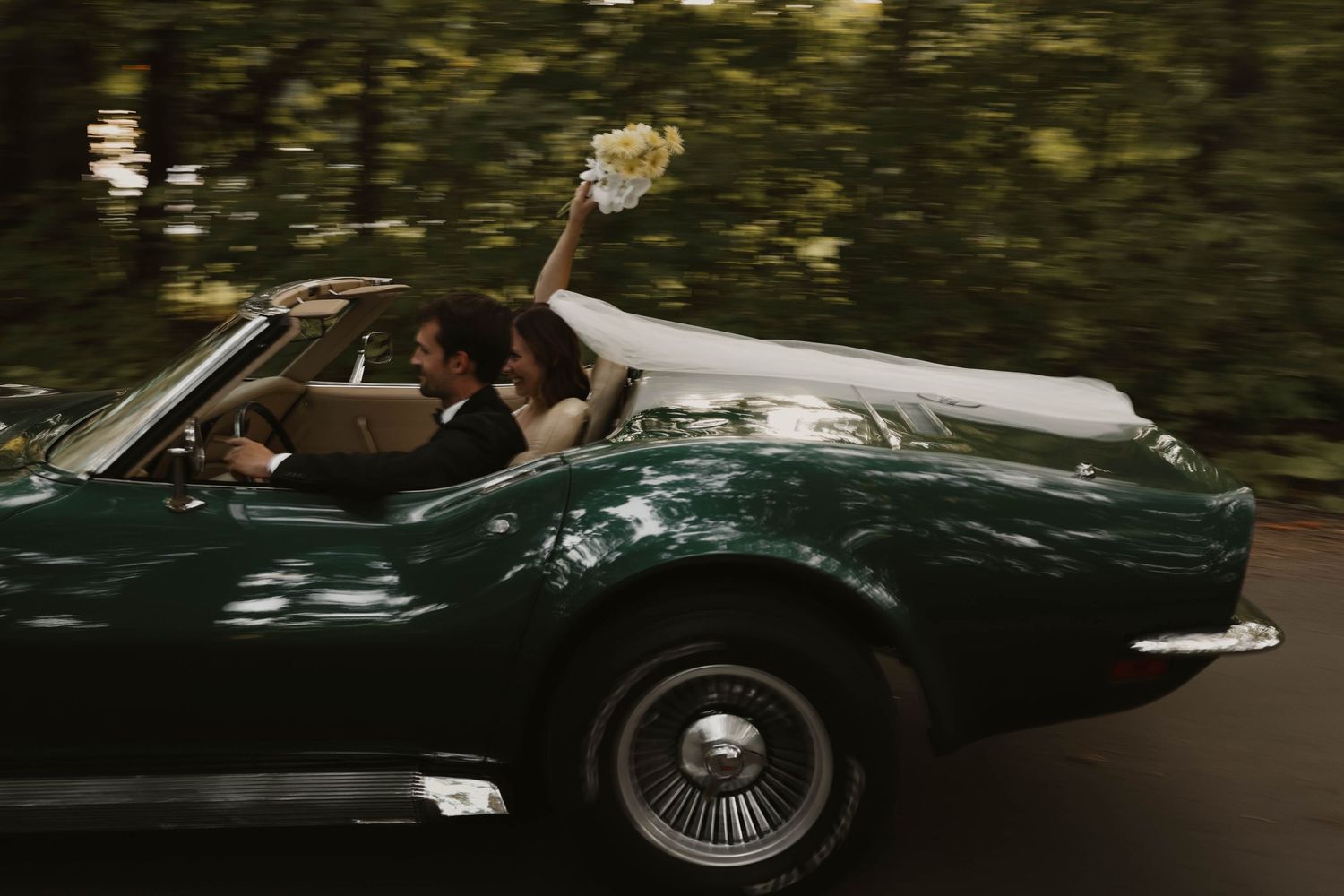 Joyful bride raising bouquet while riding in green vintage Corvette convertible with groom through forest road.