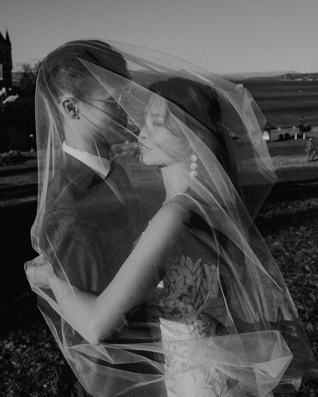 Black and white romantic photo of couple sharing a kiss under a flowing bridal veil outdoors.