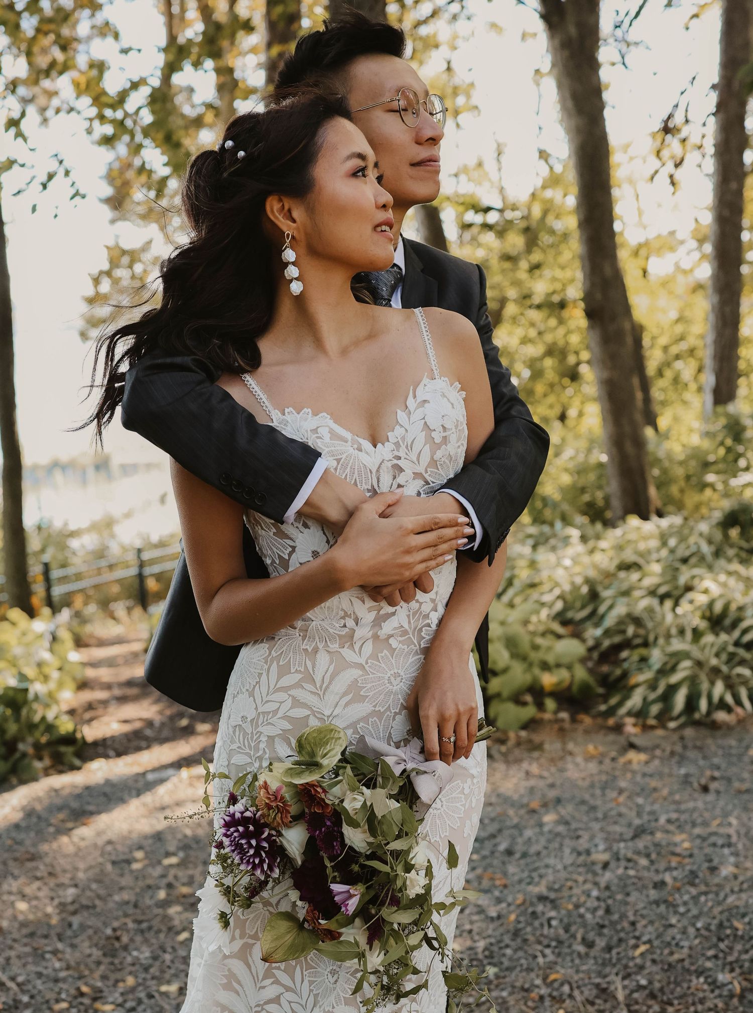Groom wraps arms around bride in lace gown holding a colorful bouquet in a wooded garden setting.