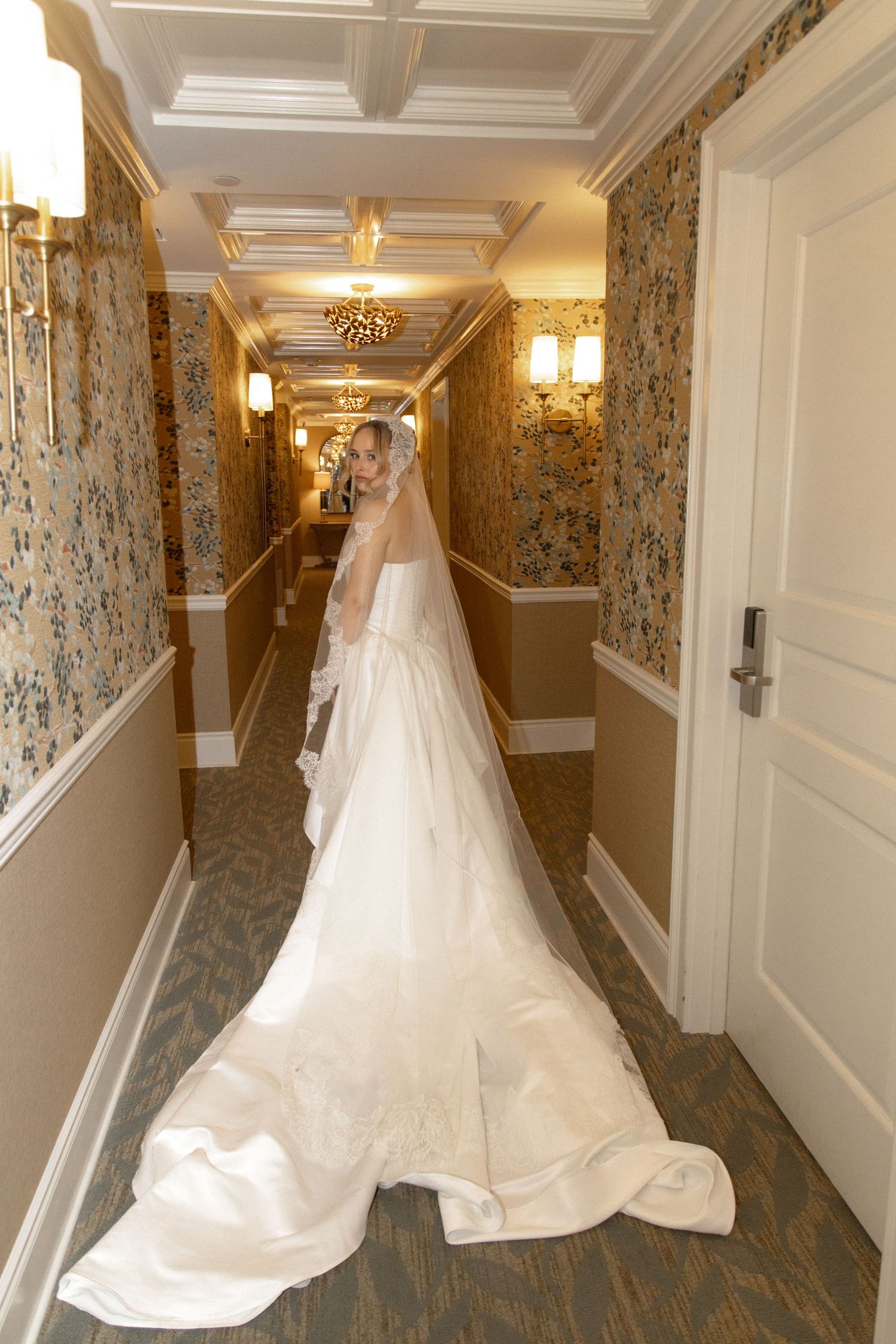 Bride in strapless white gown with long cathedral veil walking through ornate hotel corridor.