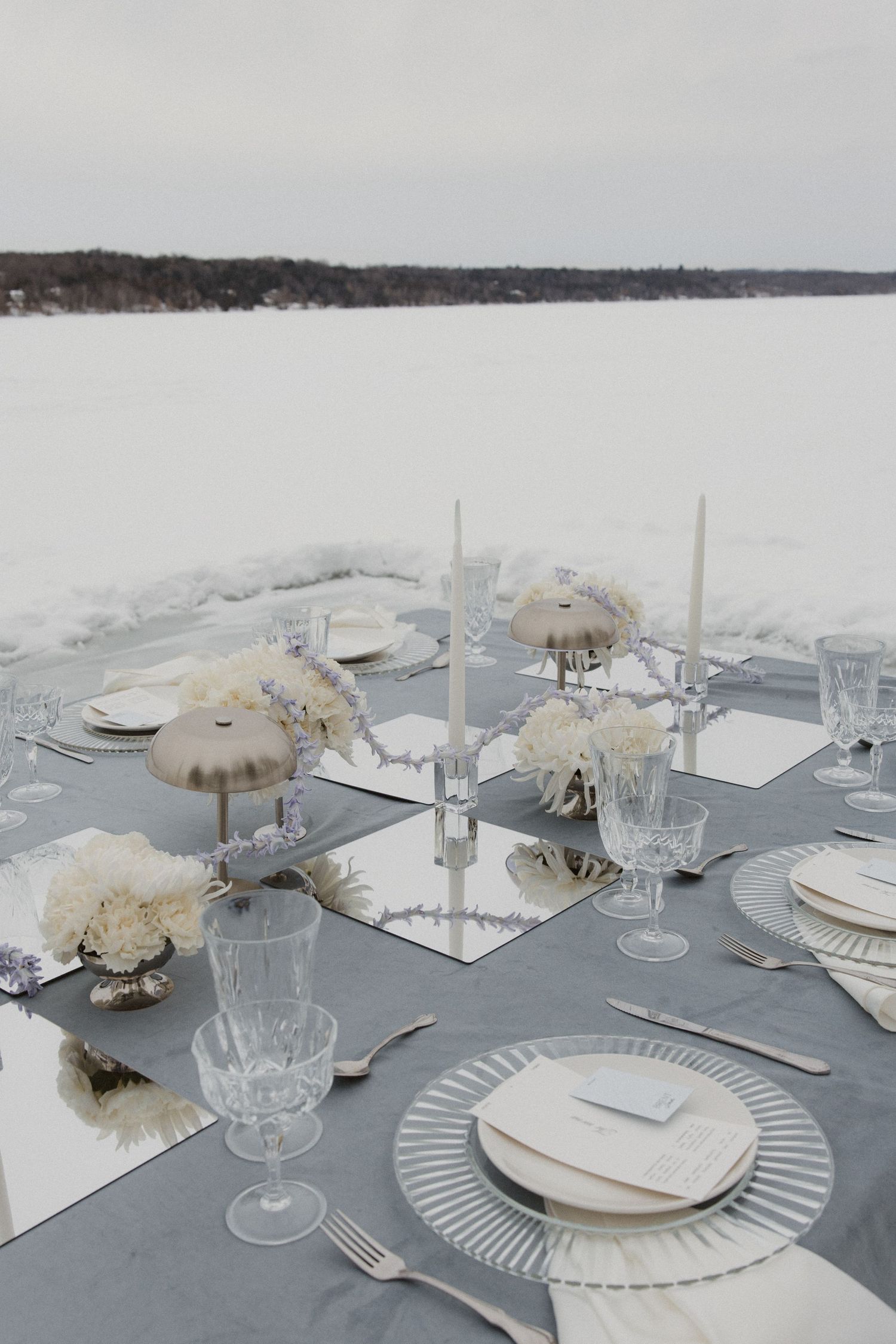 Elegant winter wedding tablescape with white florals, crystal glasses, and candles on snowy lakeside.