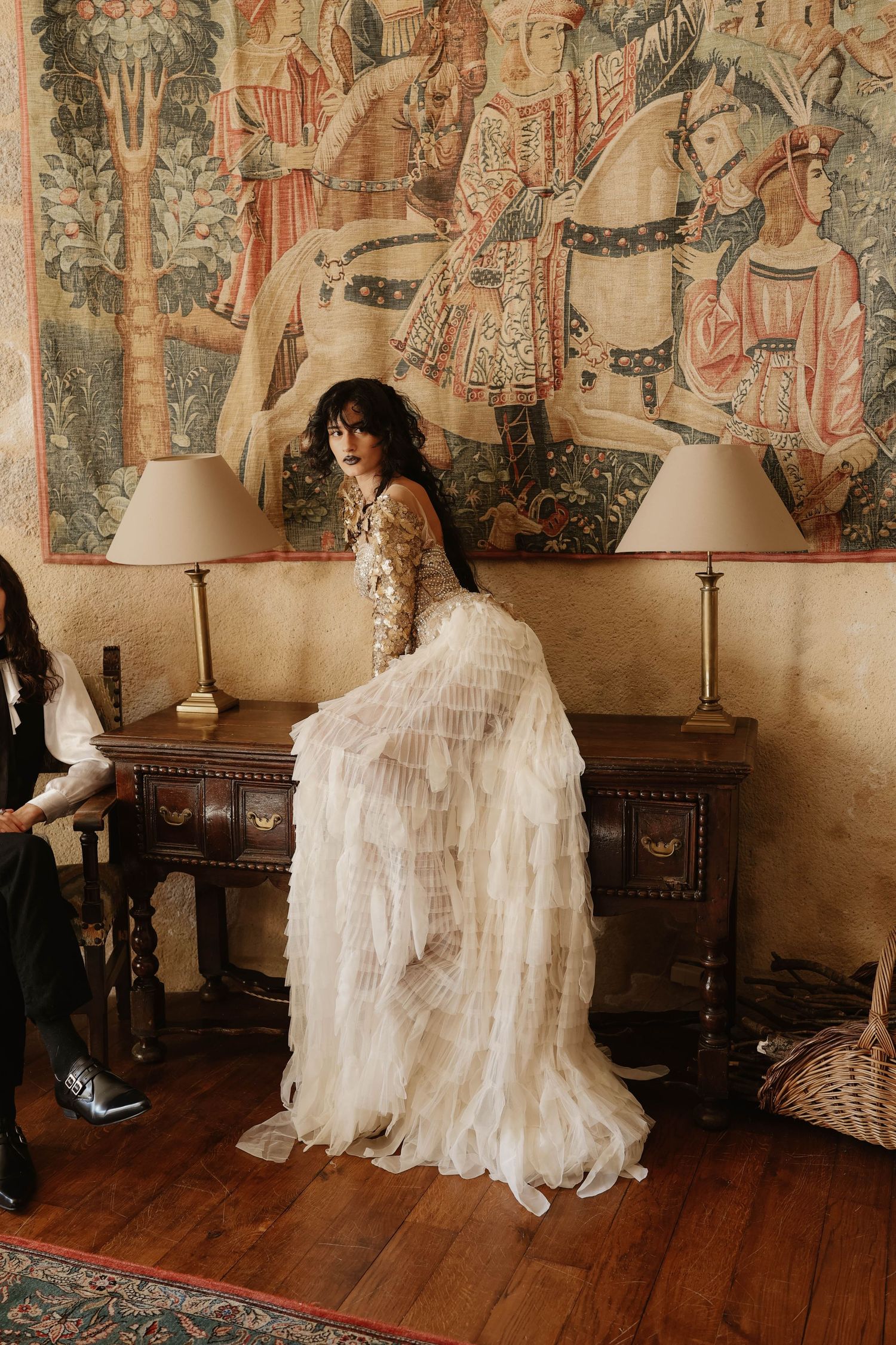 Bride in ornate beaded gown with ruffled skirt posing before a medieval tapestry.
