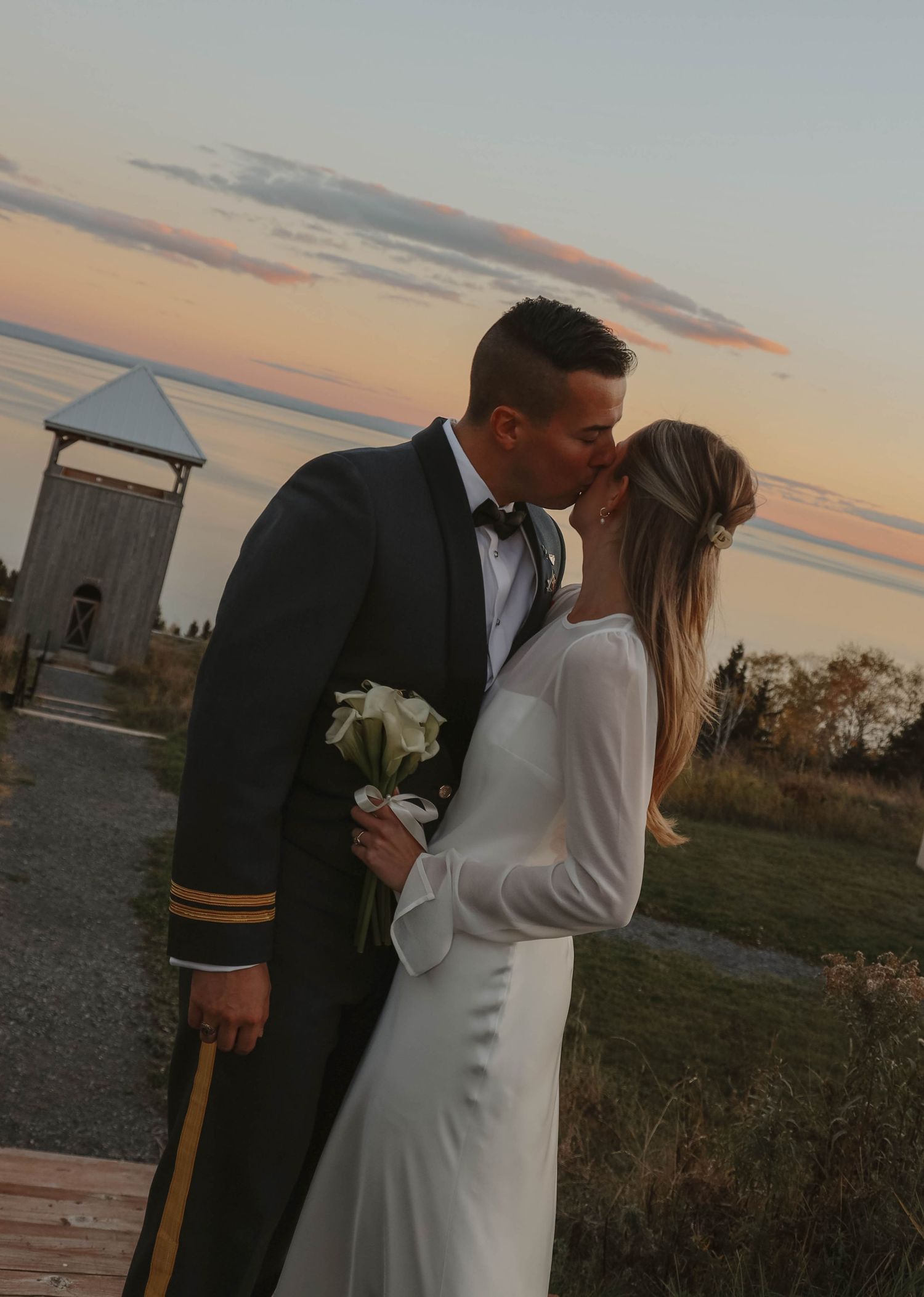 Bride and groom sharing a romantic kiss at sunset, groom wearing military dress uniform.