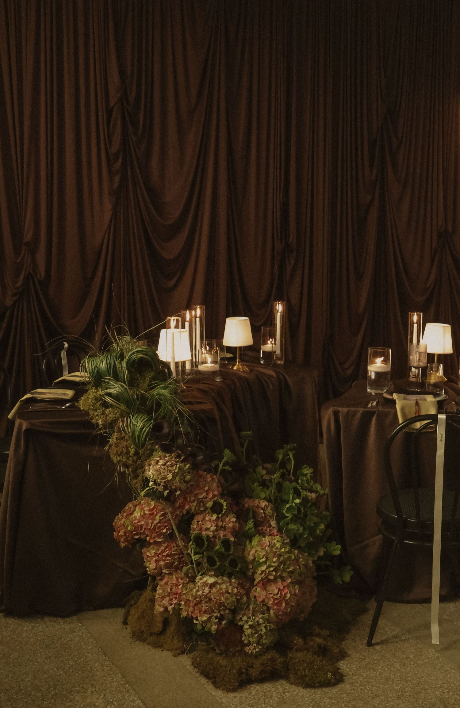 Elegant dark wedding sweetheart table with hydrangea florals, candles, and brown draped backdrop.