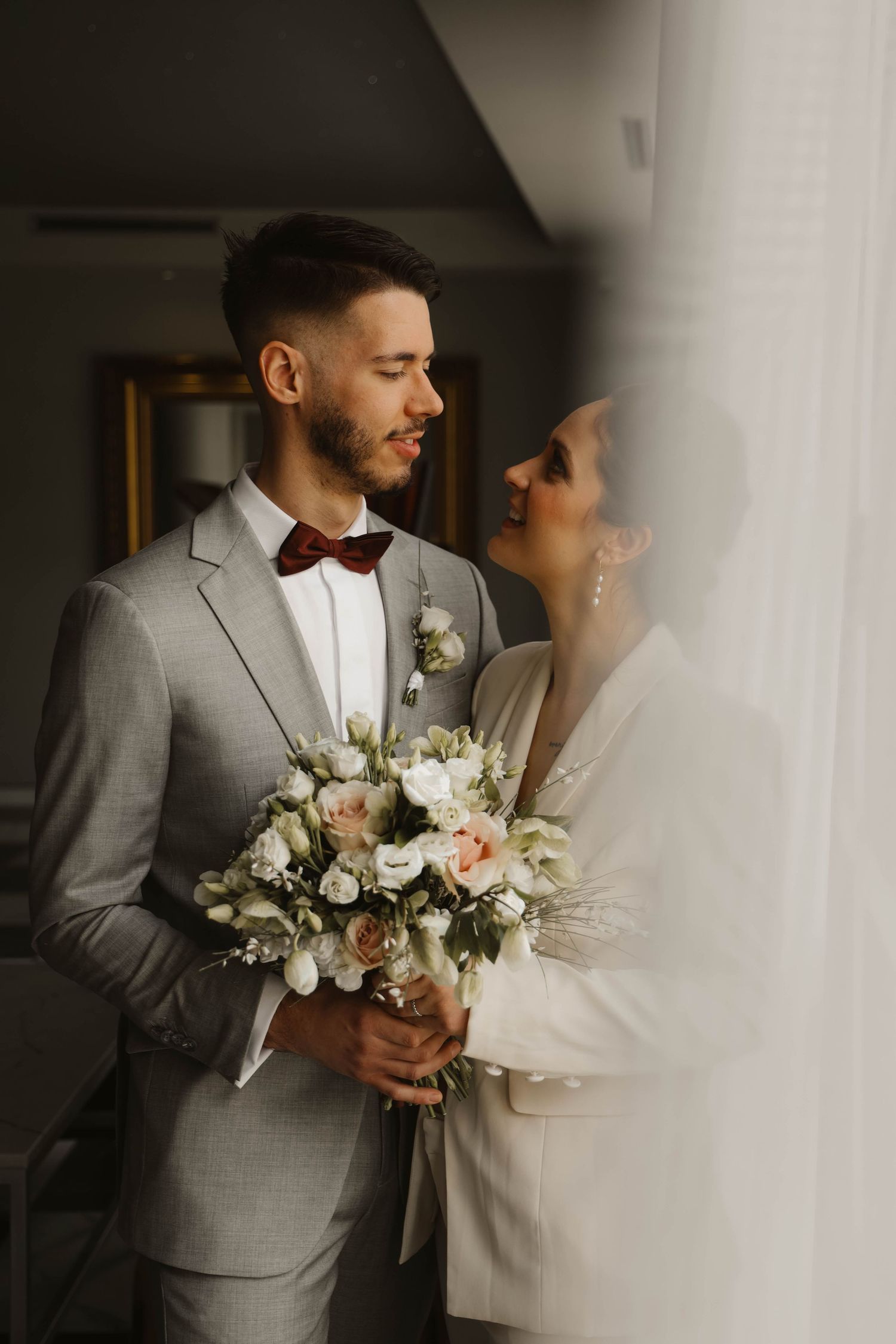 Bride and groom sharing romantic moment by window, groom in gray suit with red bow tie holding bouquet.