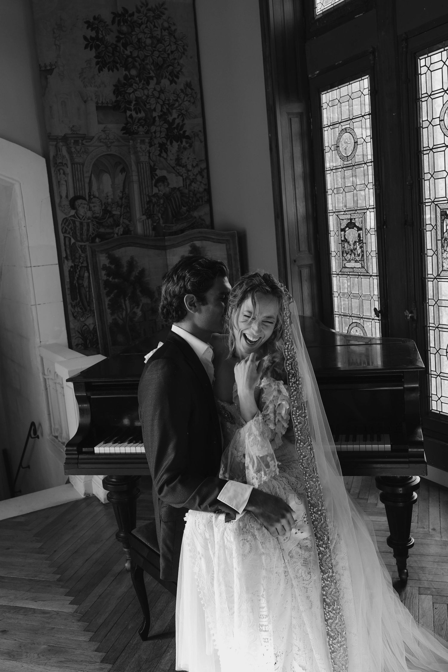 Black and white photo of bride and groom embracing near grand piano with stained glass windows backdrop.