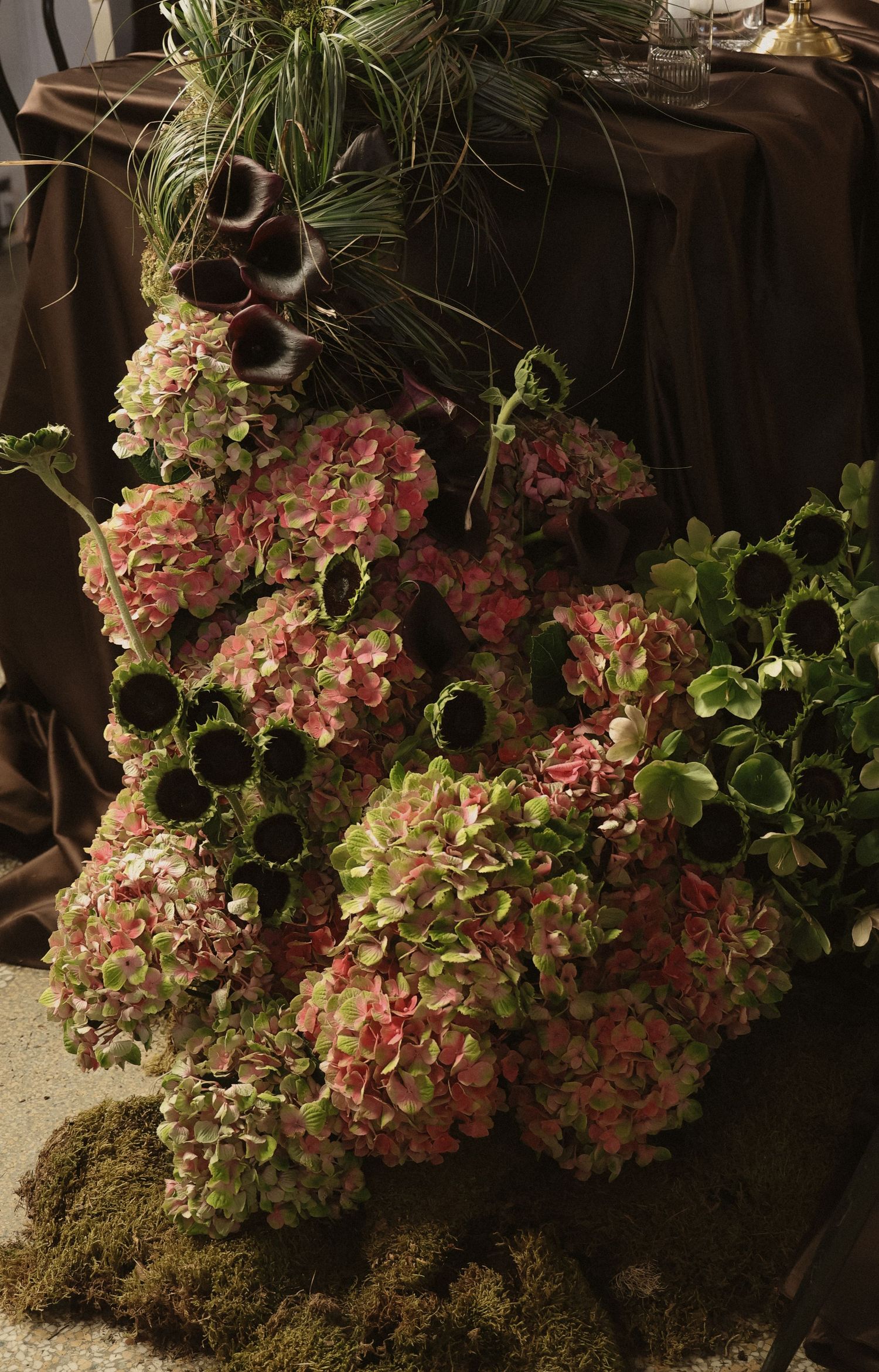 Pink and green hydrangea floral arrangement with sunflowers on moss, displayed near a draped brown table.