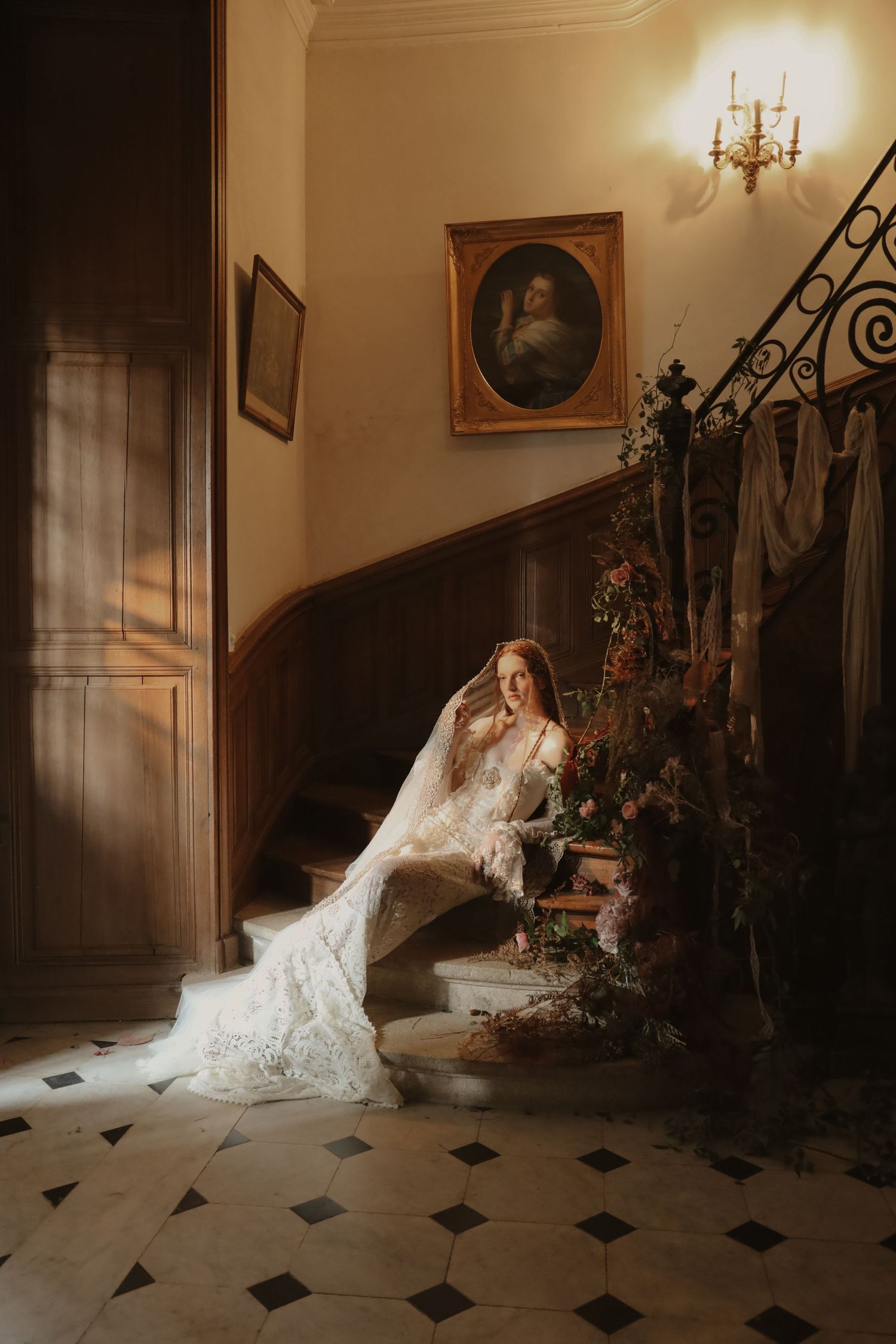 Bride in lace wedding gown seated on staircase with floral arrangement in elegant manor.