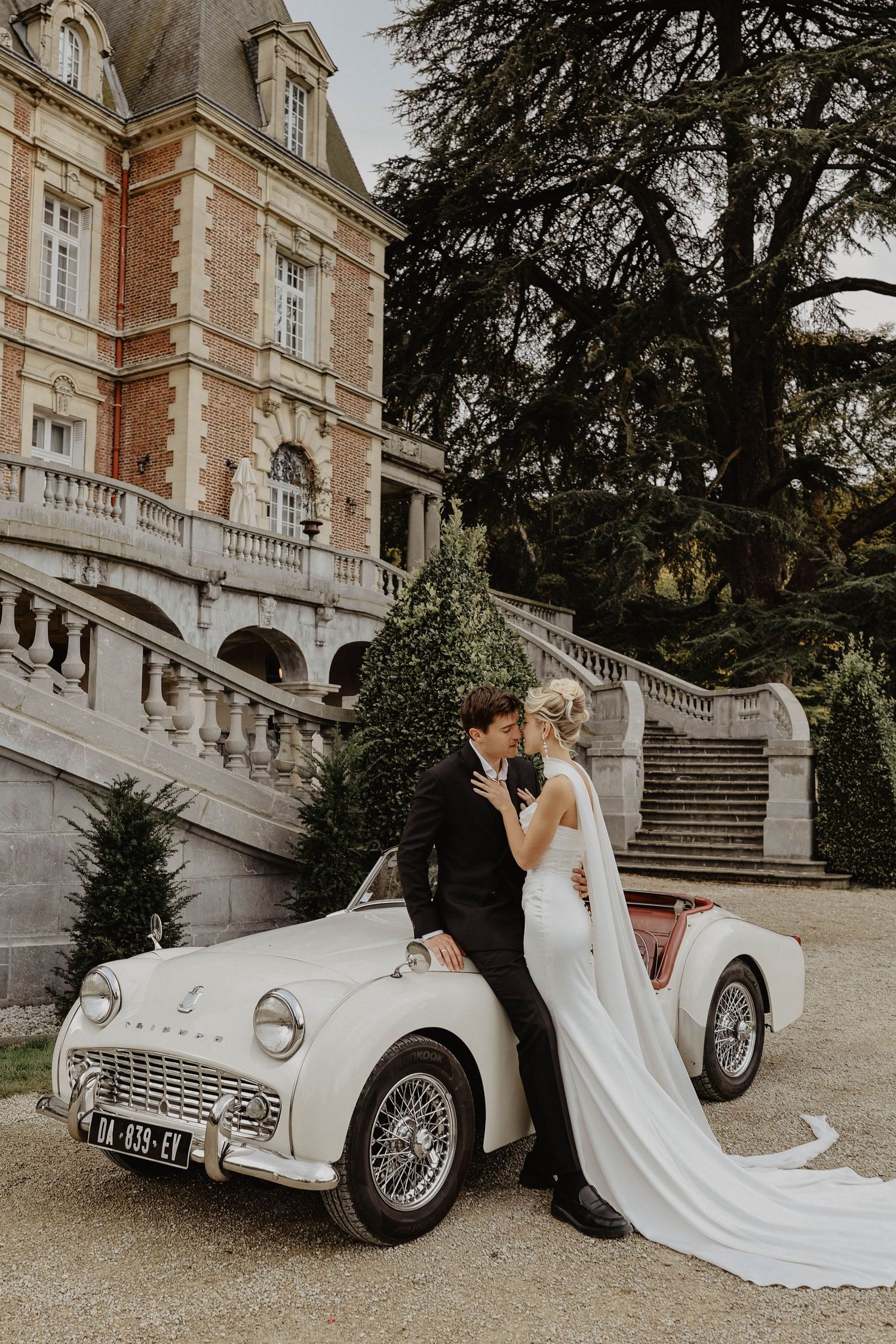 Bride and groom share a kiss leaning against a classic white Triumph TR3 car outside a French chateau.