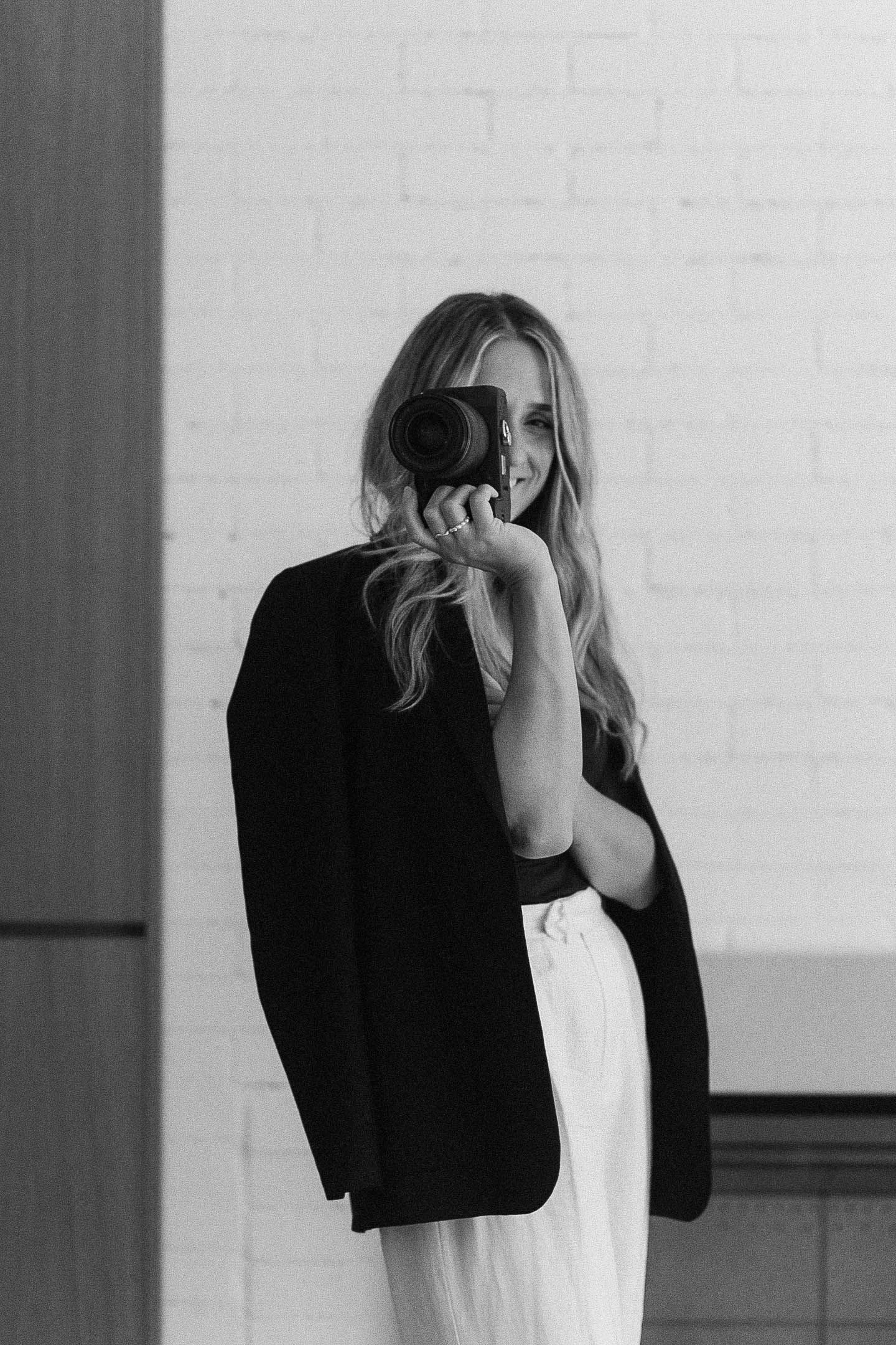 Woman in black blazer photographing with camera, turning back to viewer, black and white portrait.