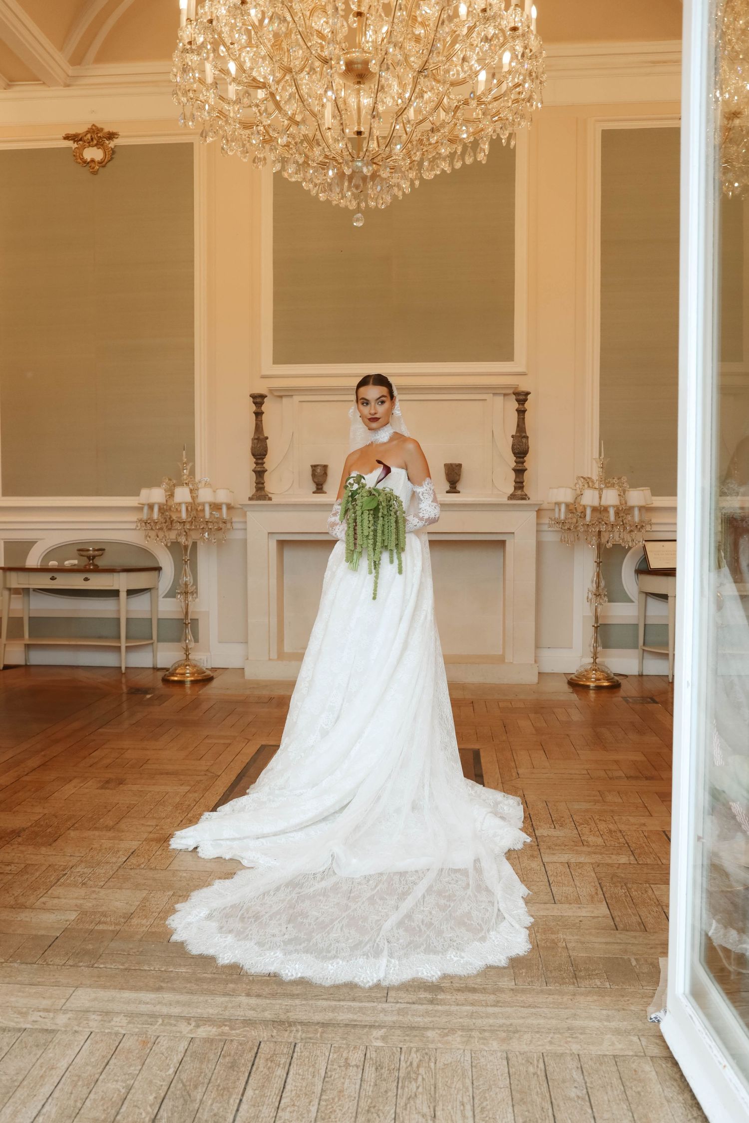 Bride in white ball gown with long train holding green bouquet in elegant chandelier ballroom.