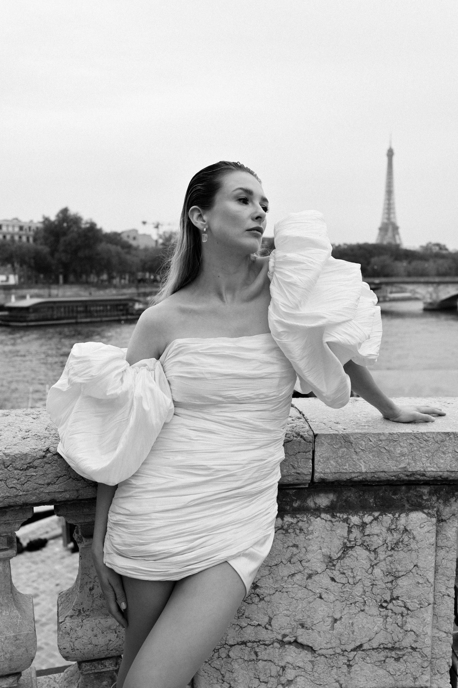 Black and white photo of woman in white ruffled dress posing on bridge with Eiffel Tower behind.