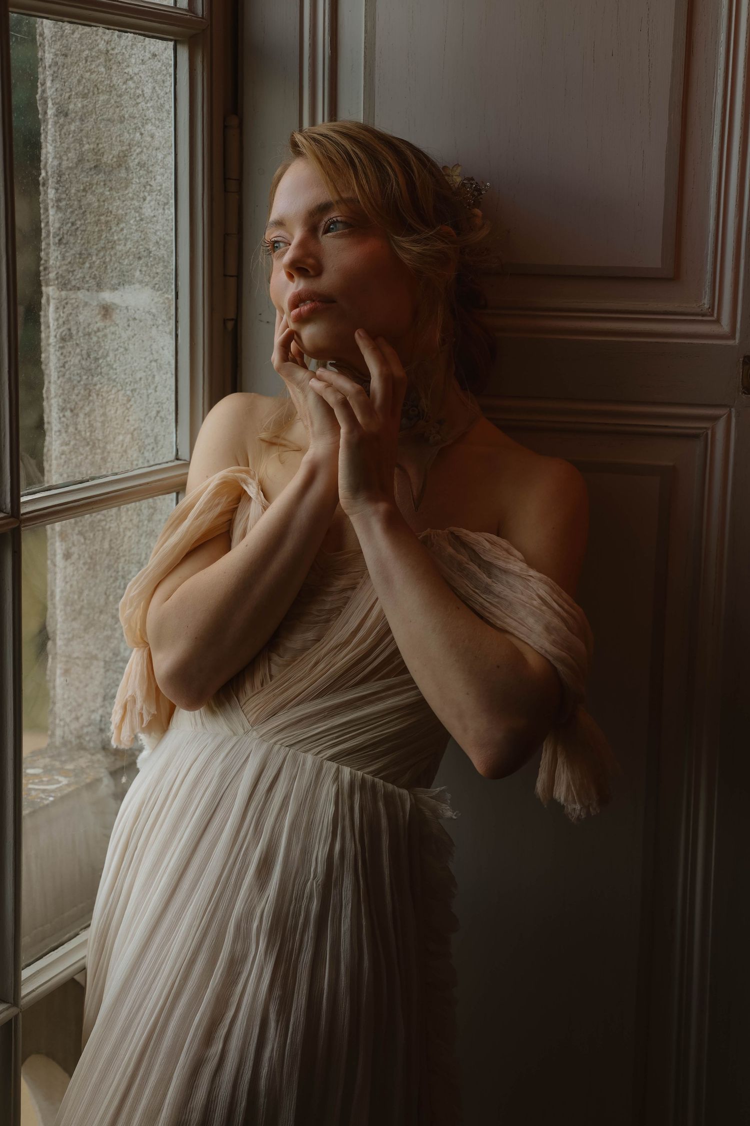 Woman in off-shoulder pleated white bridal gown stands pensively beside a window in dim interior.