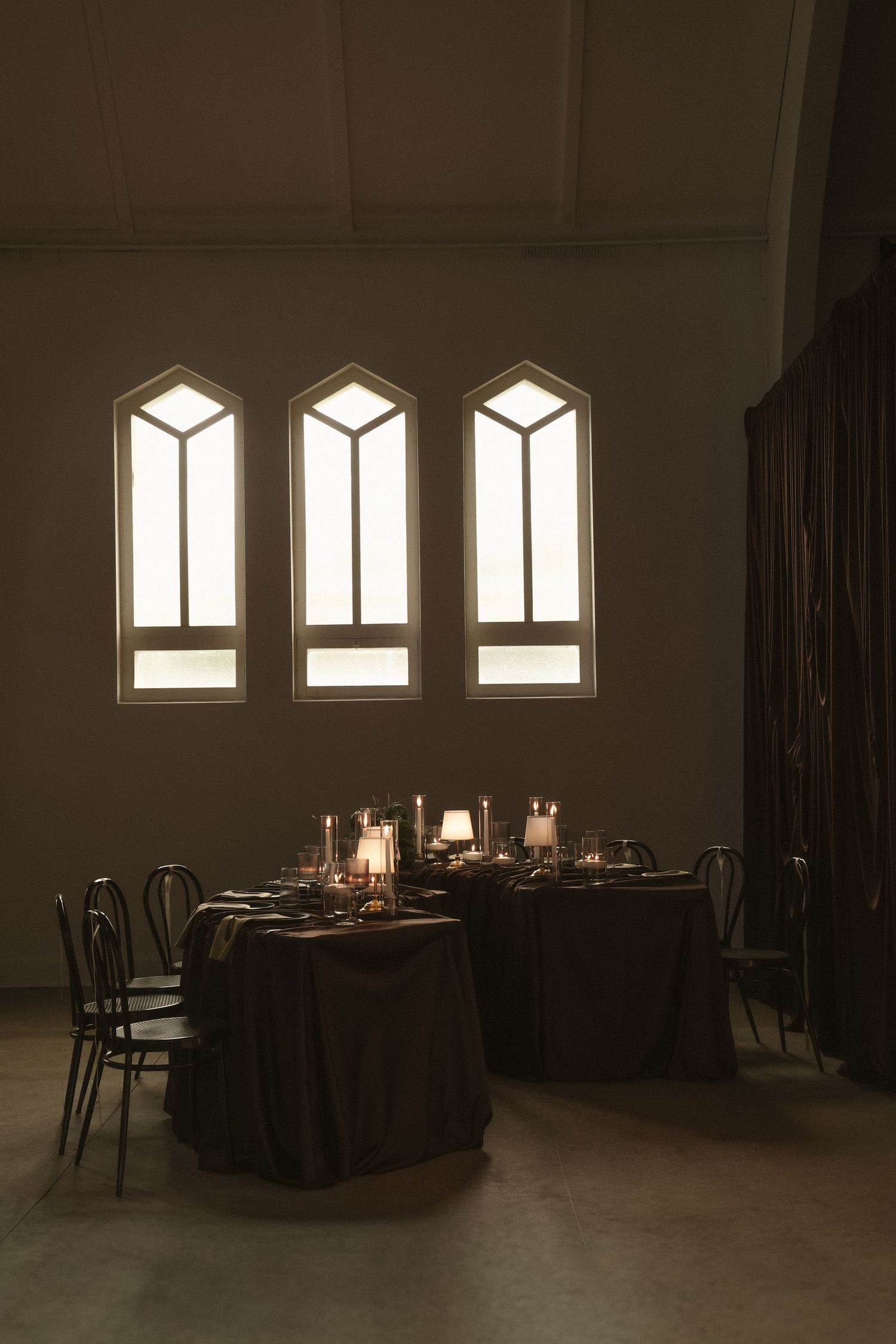 Dimly lit elegant dining room with candlelit tables and gothic arched windows casting warm light in a dark venue.