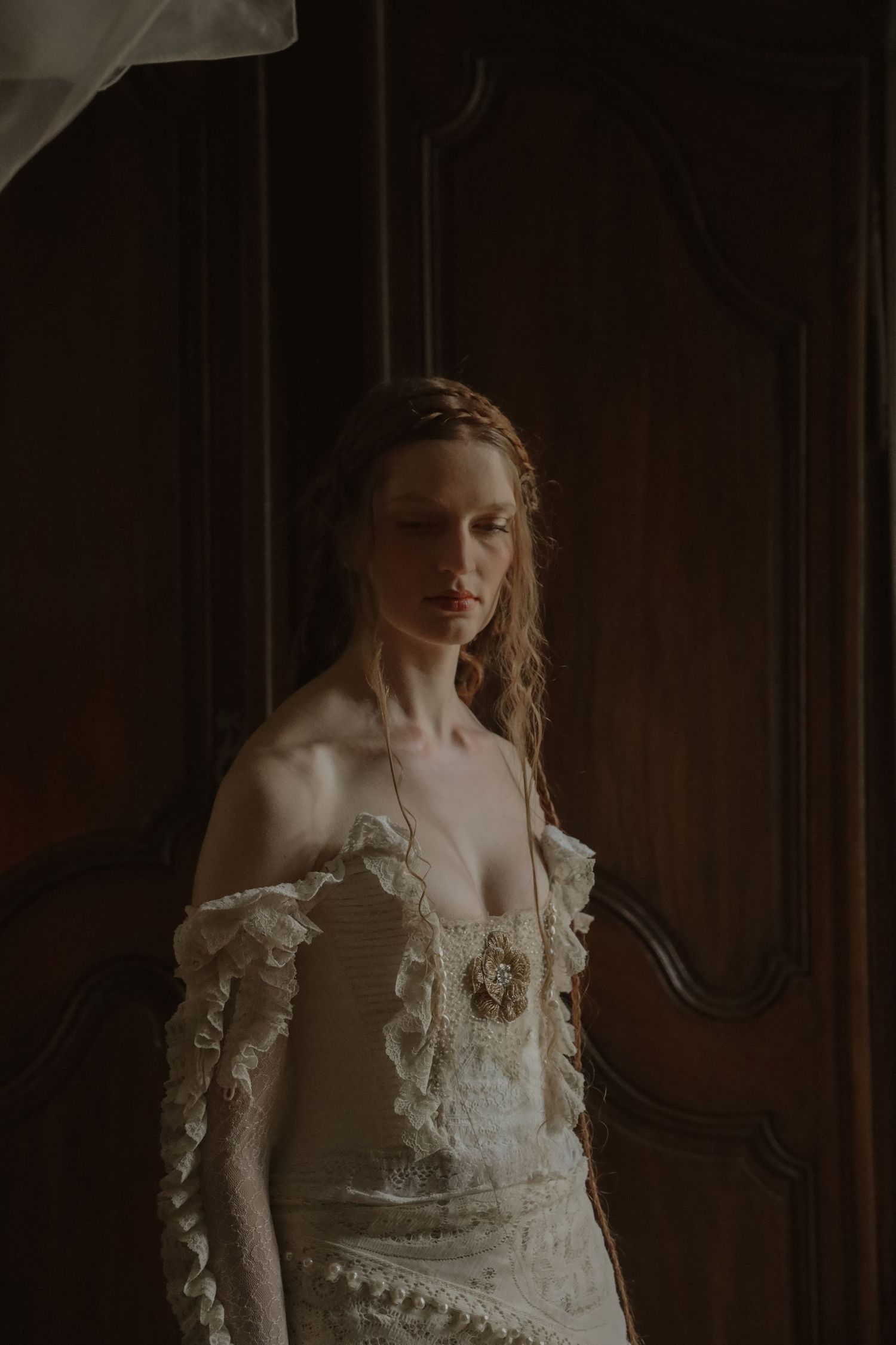 Elegant bride in off-shoulder lace wedding gown with braided hair standing by dark wood doors.