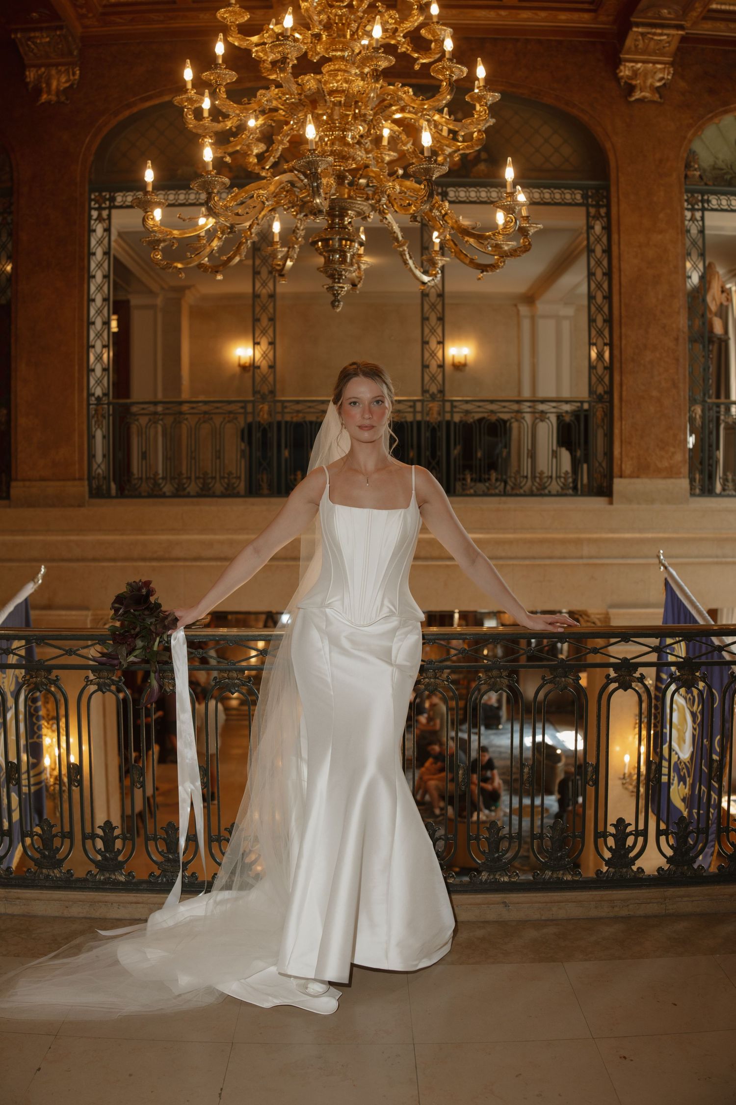 Elegant bride in white satin mermaid gown with veil stands on grand staircase under golden chandelier.