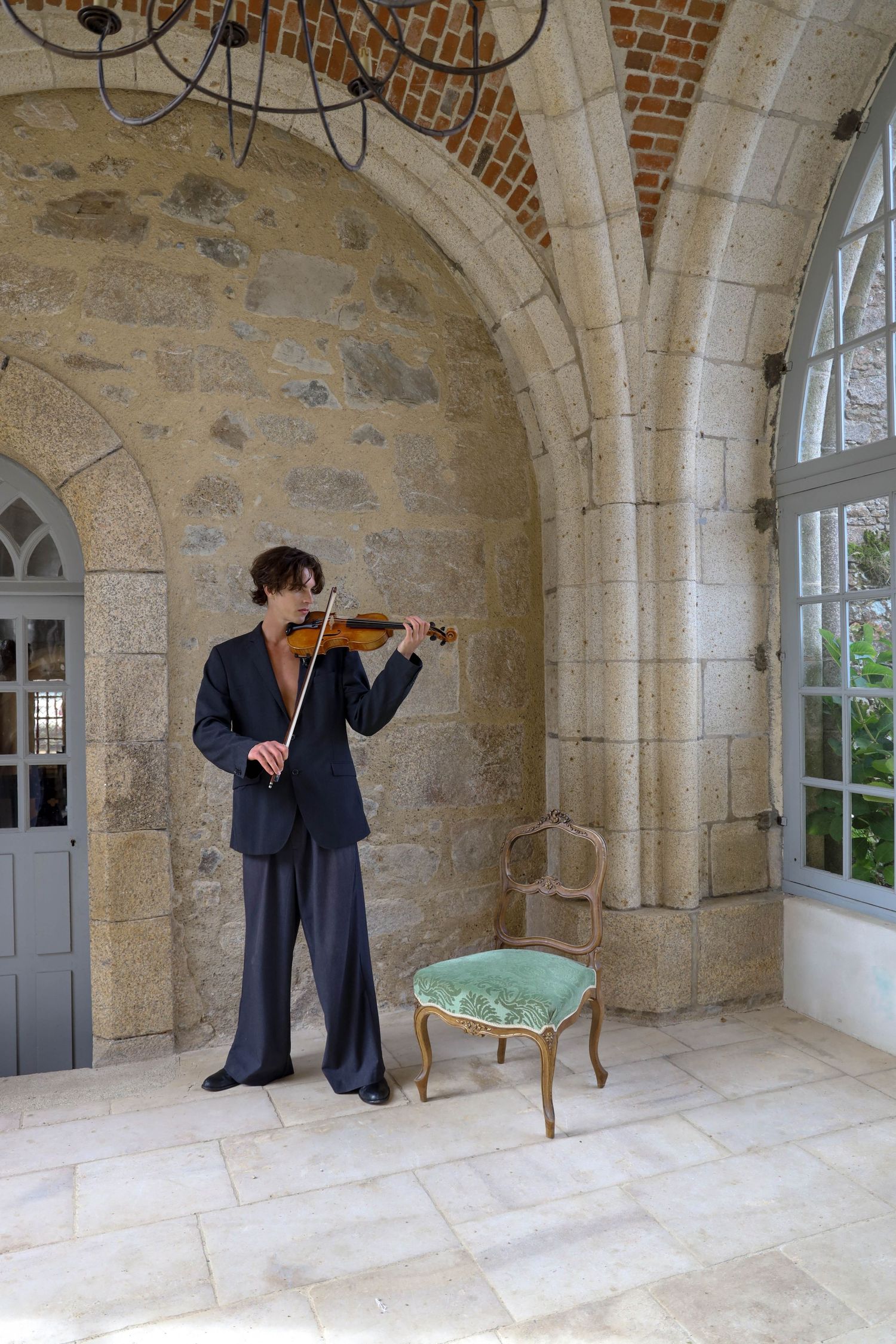 Musician in dark suit playing violin in elegant stone archway with vintage chair.