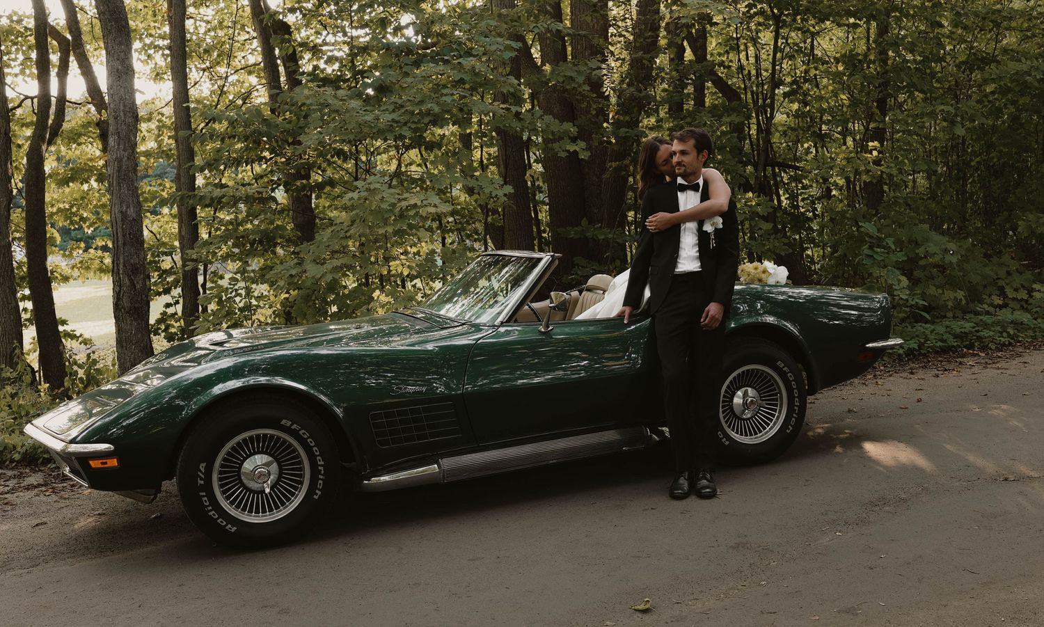 Couple posing beside a green Corvette convertible surrounded by lush forest trees.