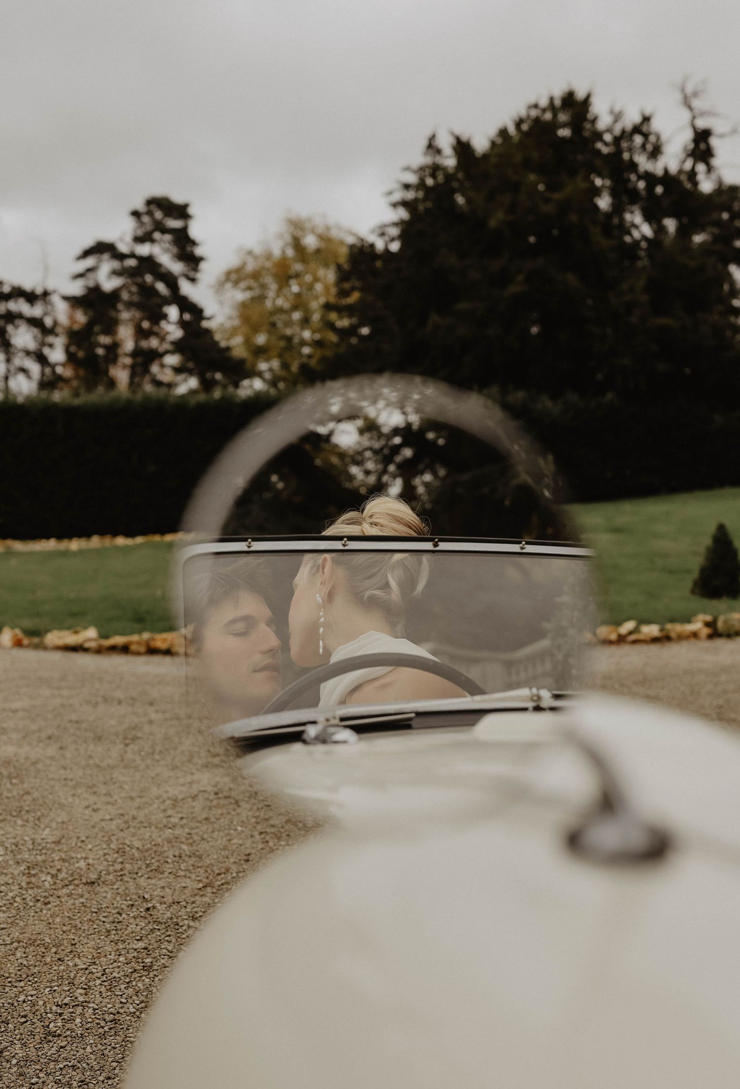 Artistic wedding photo of bride and groom kissing inside a vintage white convertible car, shot through windshield.